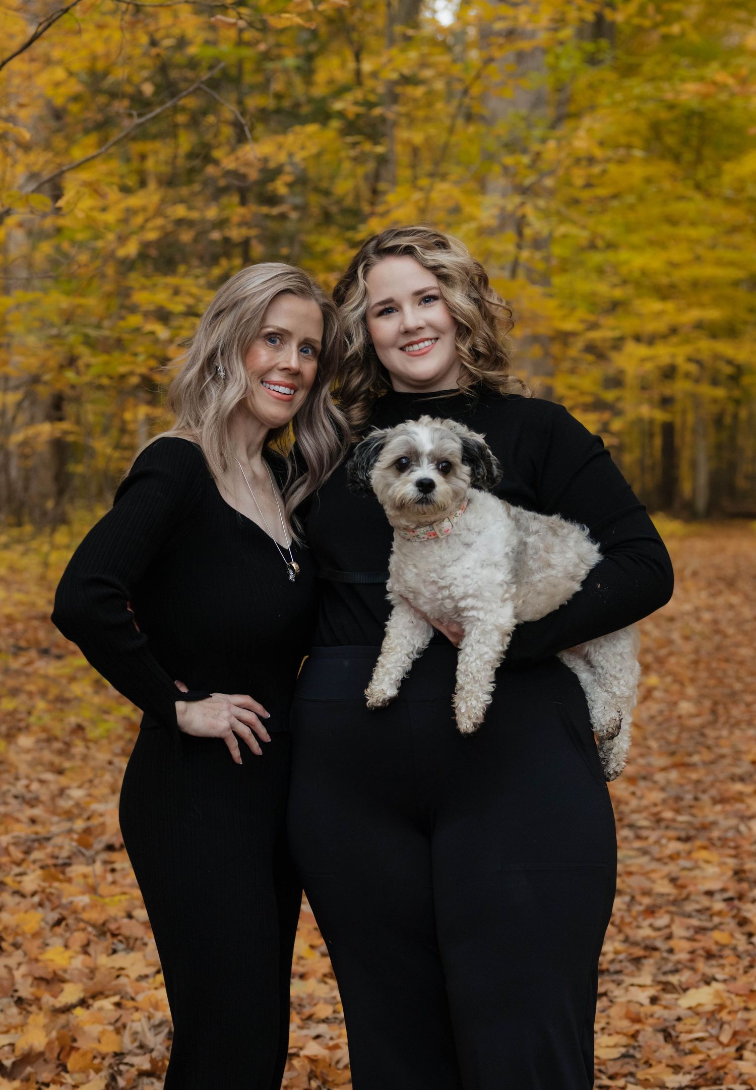 Two women in black outfits holding a fluffy dog among golden autumn foliage background.