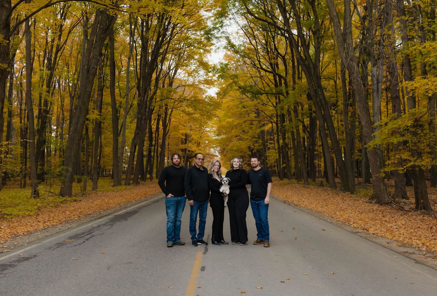 Family group of five standing on a tree-lined road surrounded by golden fall foliage.
