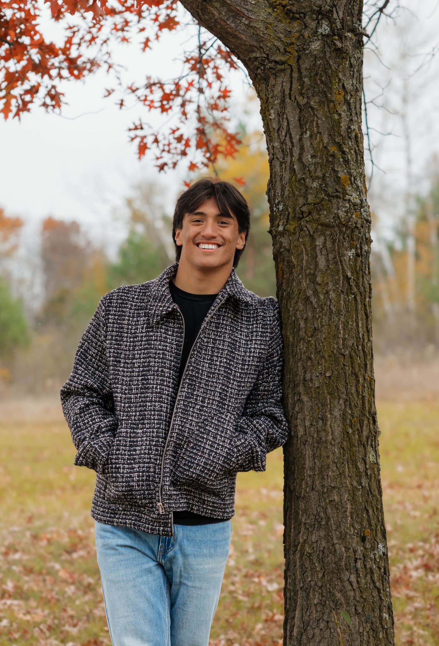 Young man in plaid jacket leaning against tree with autumn foliage in outdoor fall photo session.