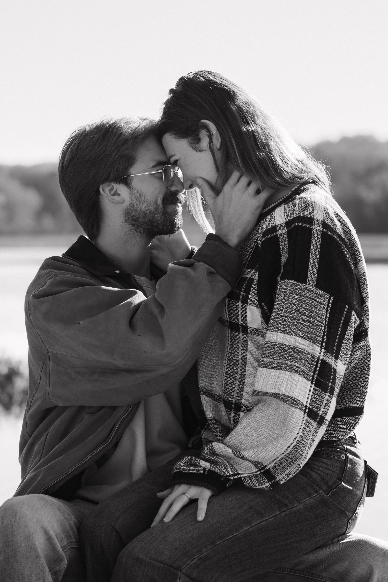 Black and white romantic photo of couple almost kissing by a lake in autumn.