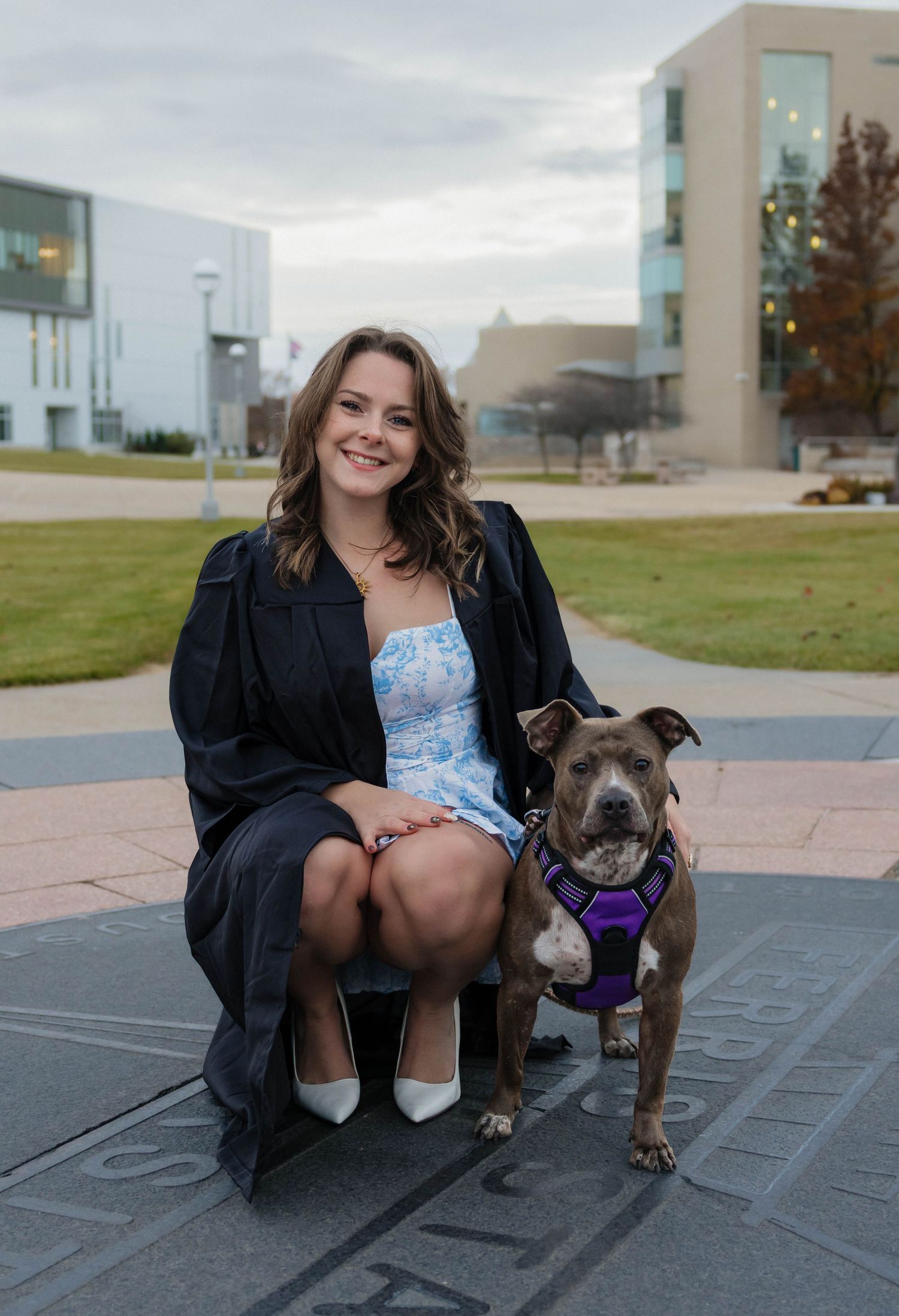 Smiling female graduate in cap and gown posing with a gray pit bull dog on a university campus.