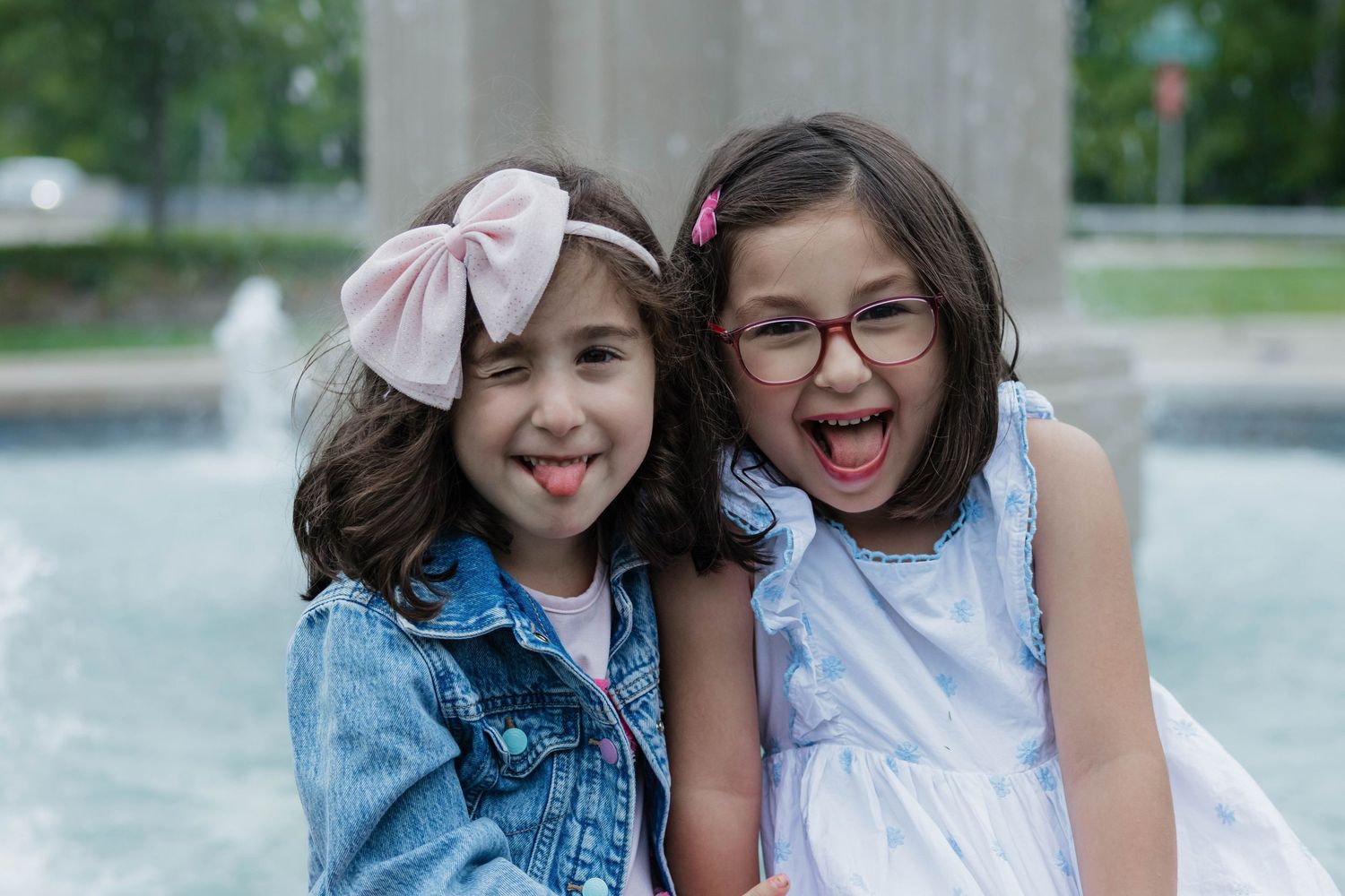 Two young girls making silly faces together in front of outdoor fountain, one wearing denim jacket.