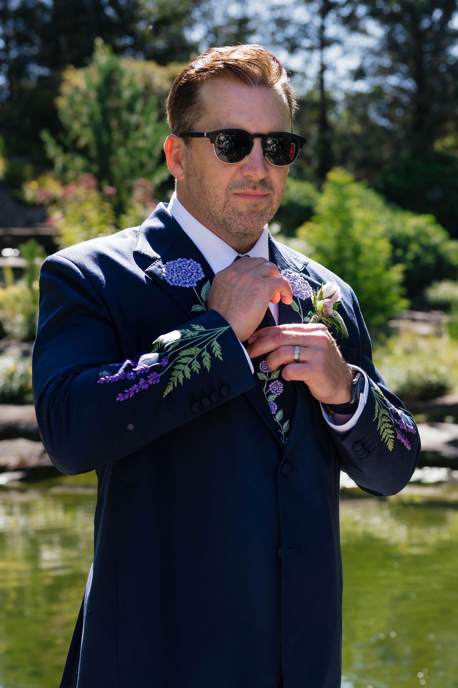 Stylish groom in navy floral-embroidered suit adjusts boutonniere while wearing sunglasses in outdoor garden setting.