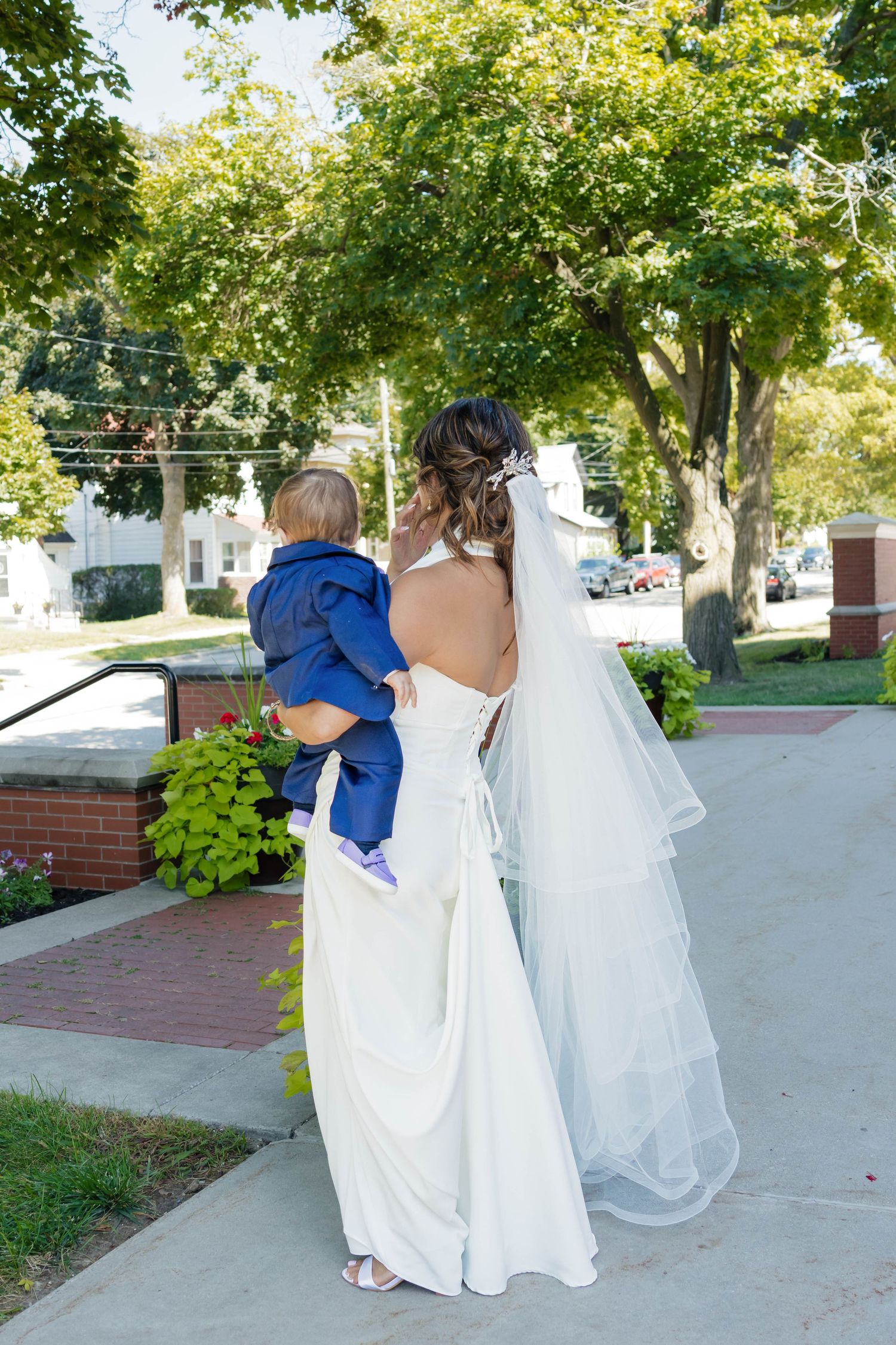 Bride in backless white gown with veil carrying a baby in a blue suit outdoors.