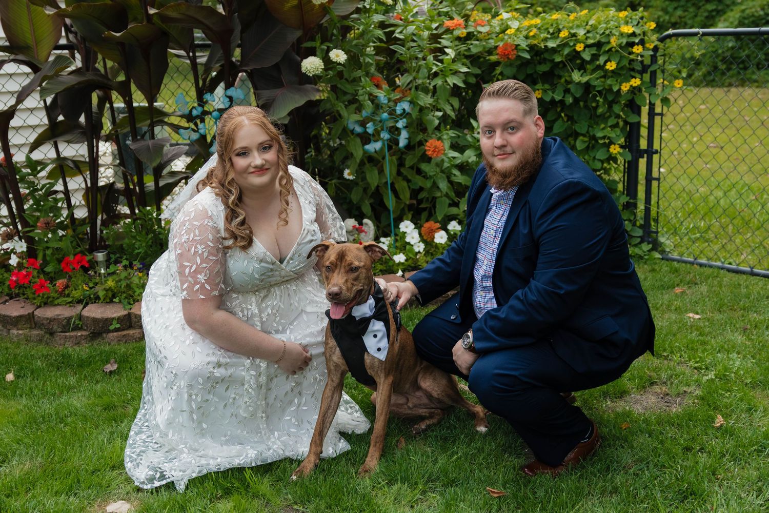 Wedding couple posing with brown dog in tuxedo in garden with red flowers.