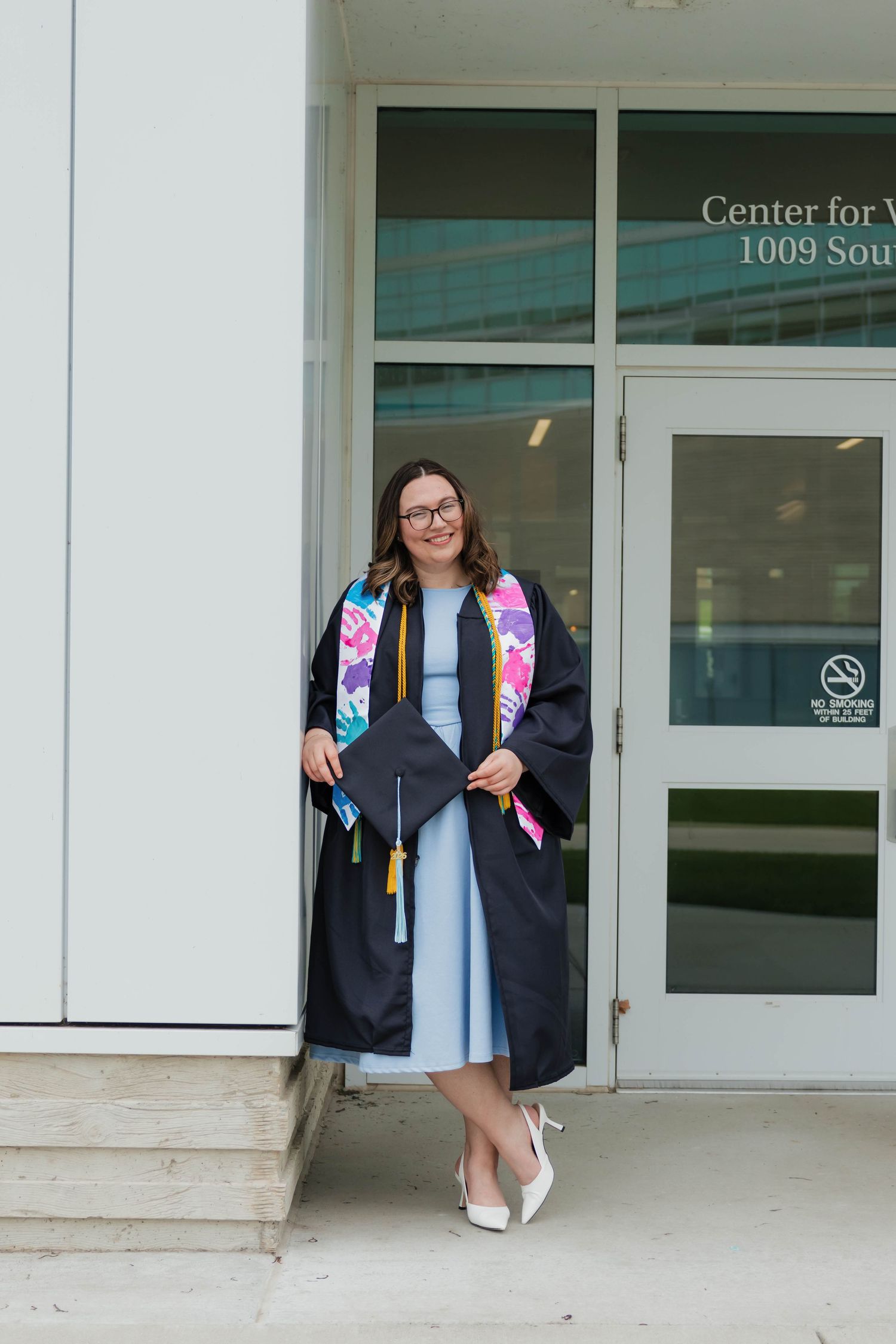 Smiling graduate in blue dress and black gown with colorful stole posing outside Center for Visual Arts.
