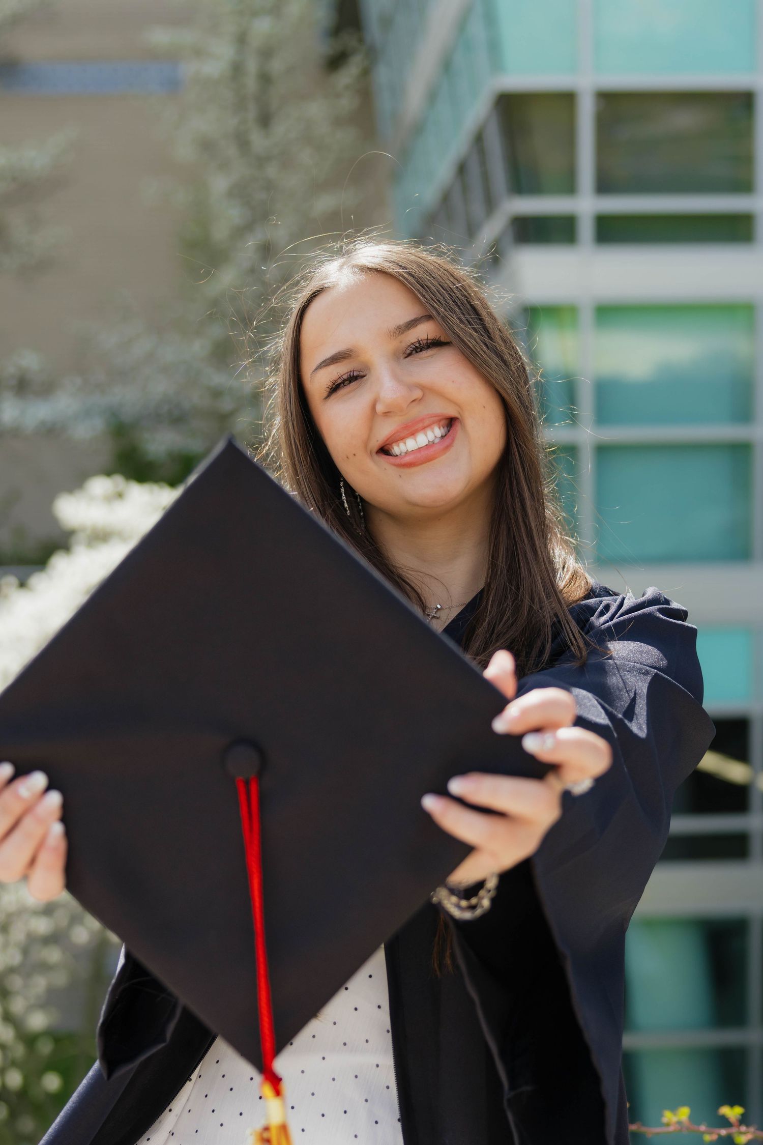 Joyful female graduate in cap and gown holding mortarboard toward camera outdoors.