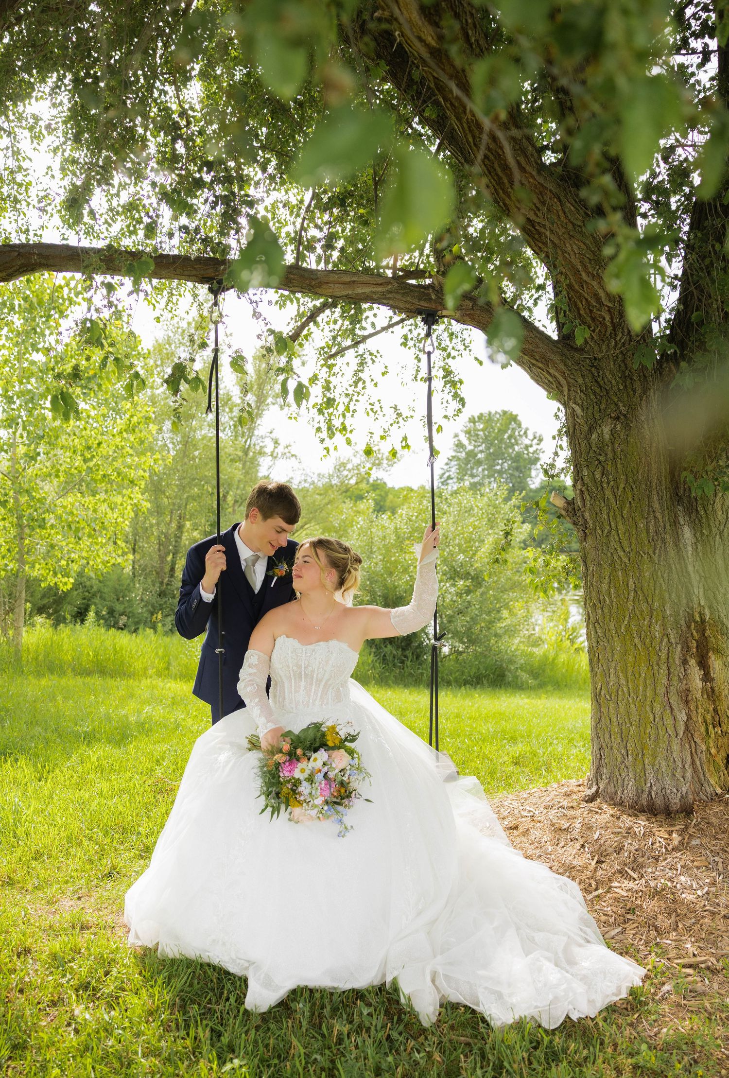 Bride and groom share a romantic moment on a tree swing, bride holding a colorful bouquet in a lush garden.
