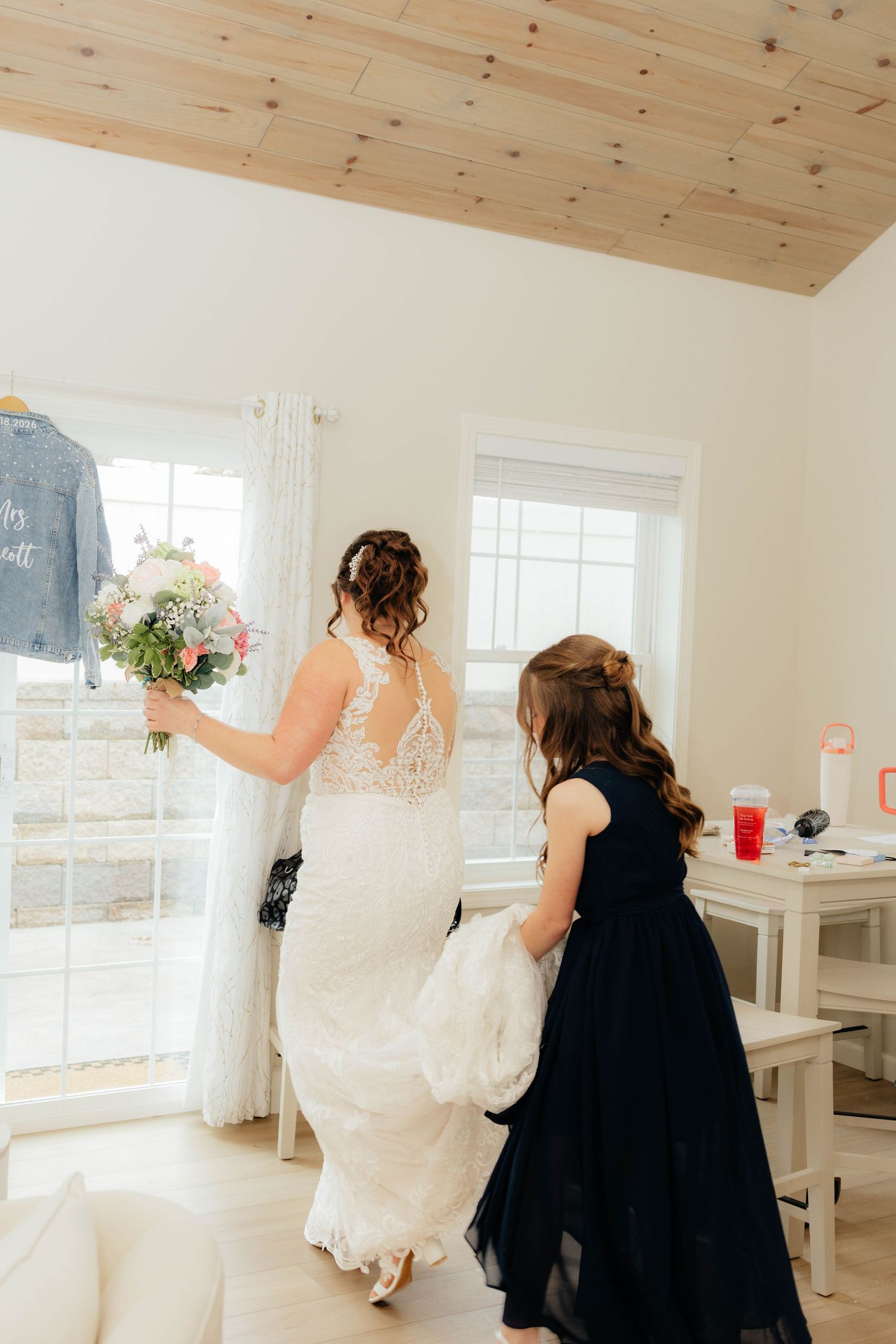 Bride in lace wedding gown holds floral bouquet while flower girl in navy dress helps carry her train indoors.