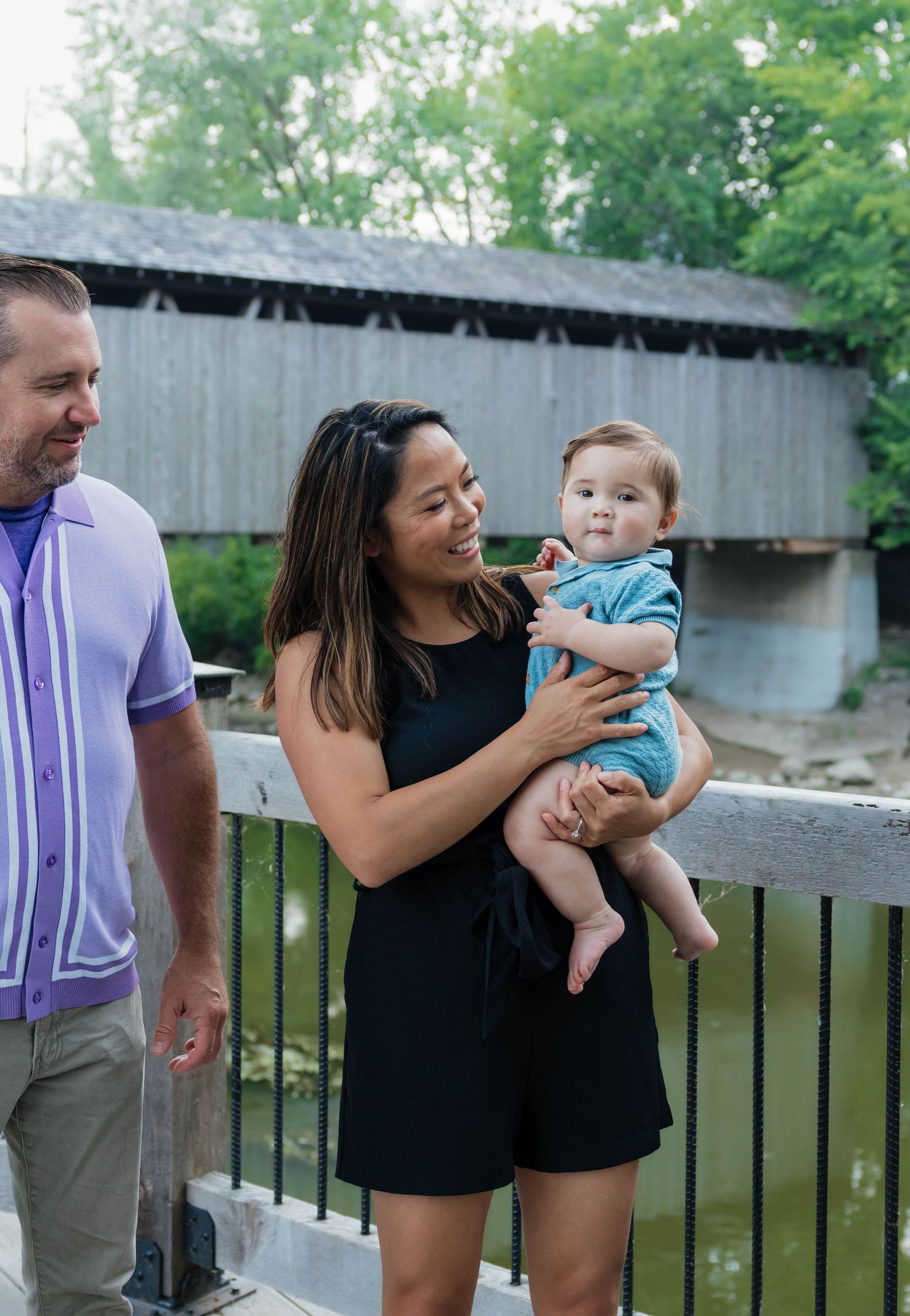 Mother holding baby boy while father smiles beside a covered bridge.