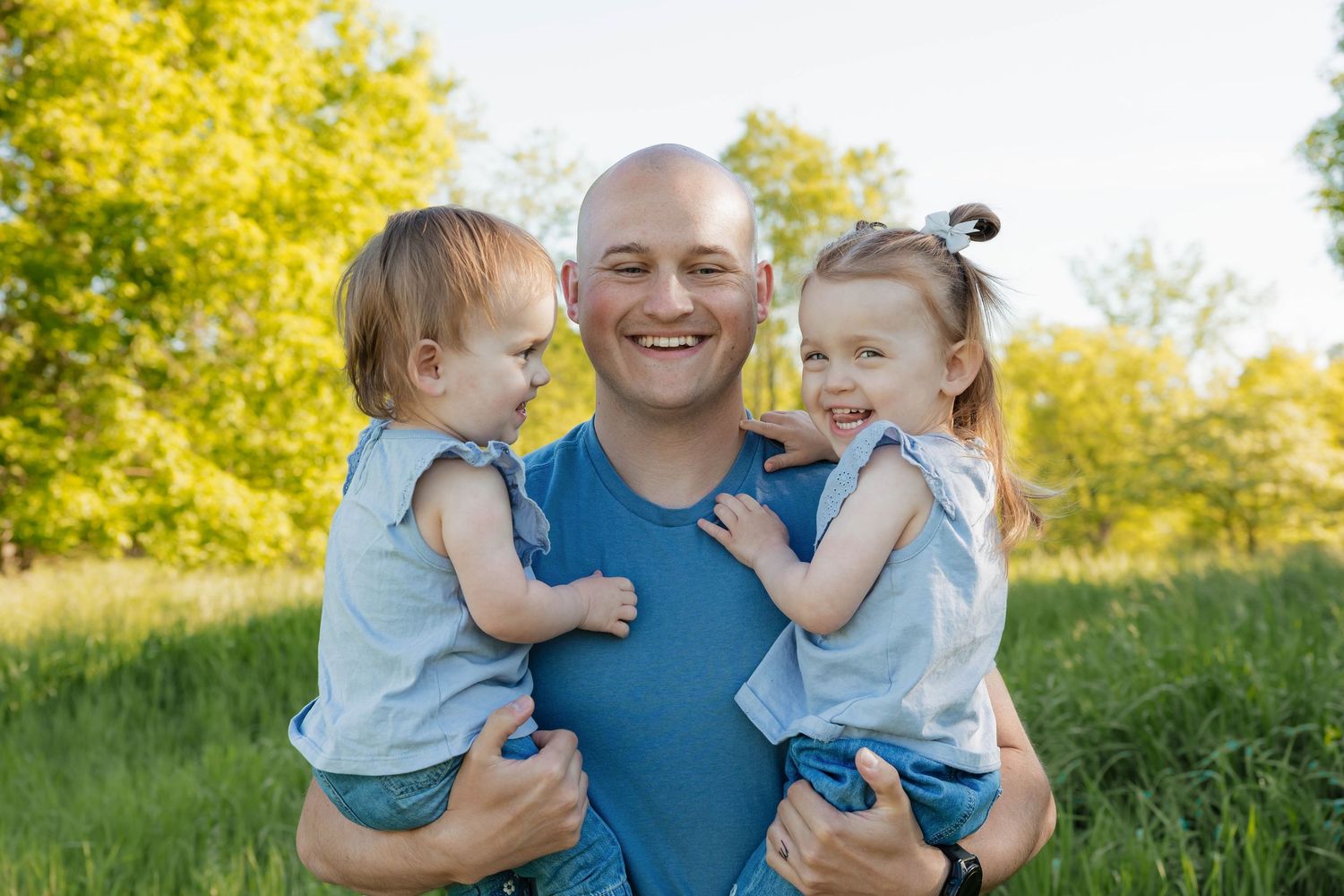 Smiling bald father holds two young daughters in blue outfits in sunny green field.