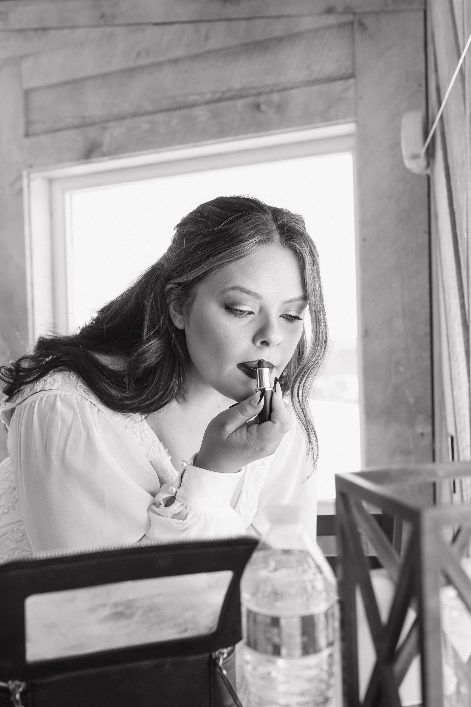 Black and white photo of a bride applying lipstick while seated near a window before her wedding.