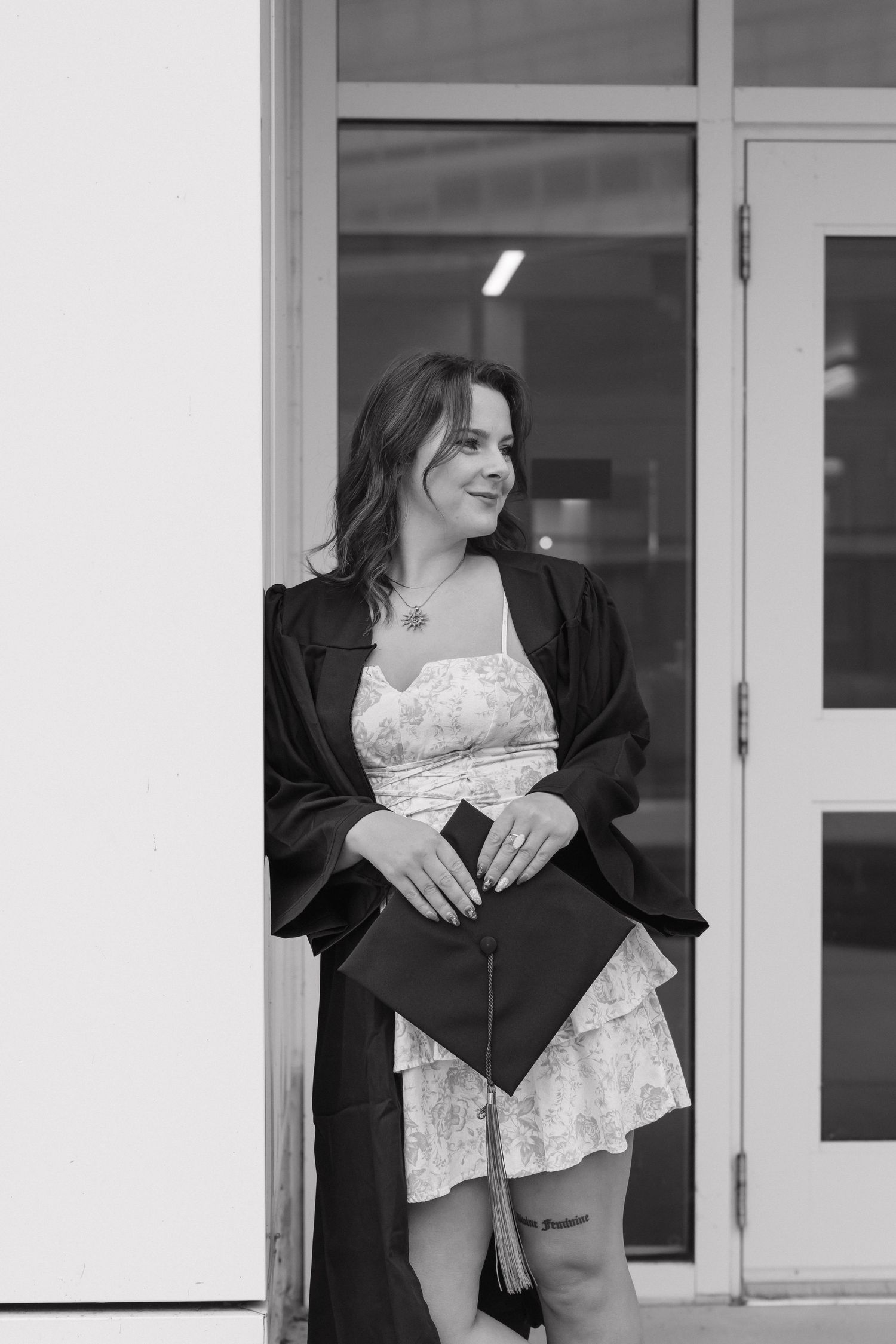 Black and white photo of a young woman in graduation gown holding cap while leaning against a building.