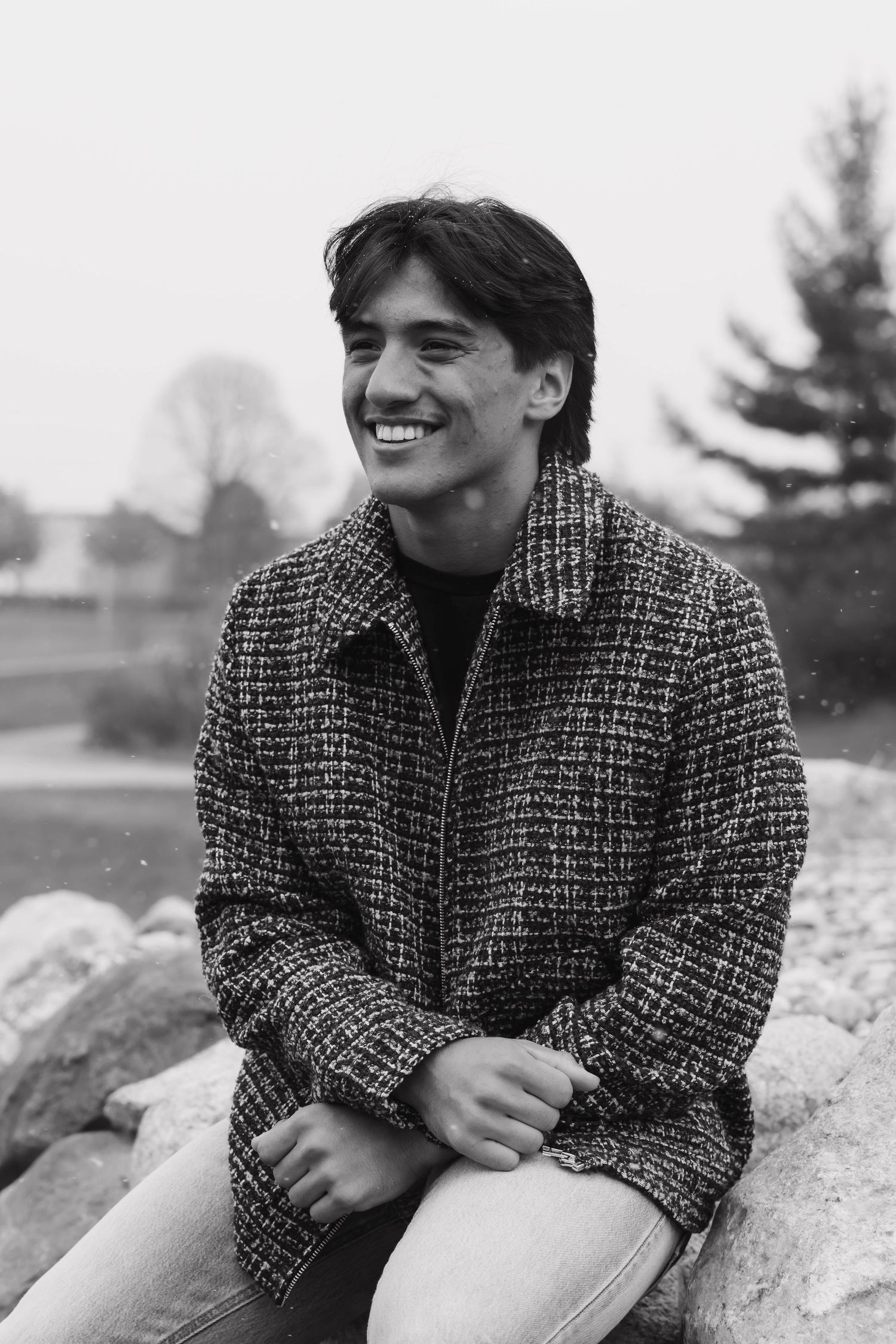 Black and white portrait of smiling young man in textured jacket sitting on rocks outdoors.