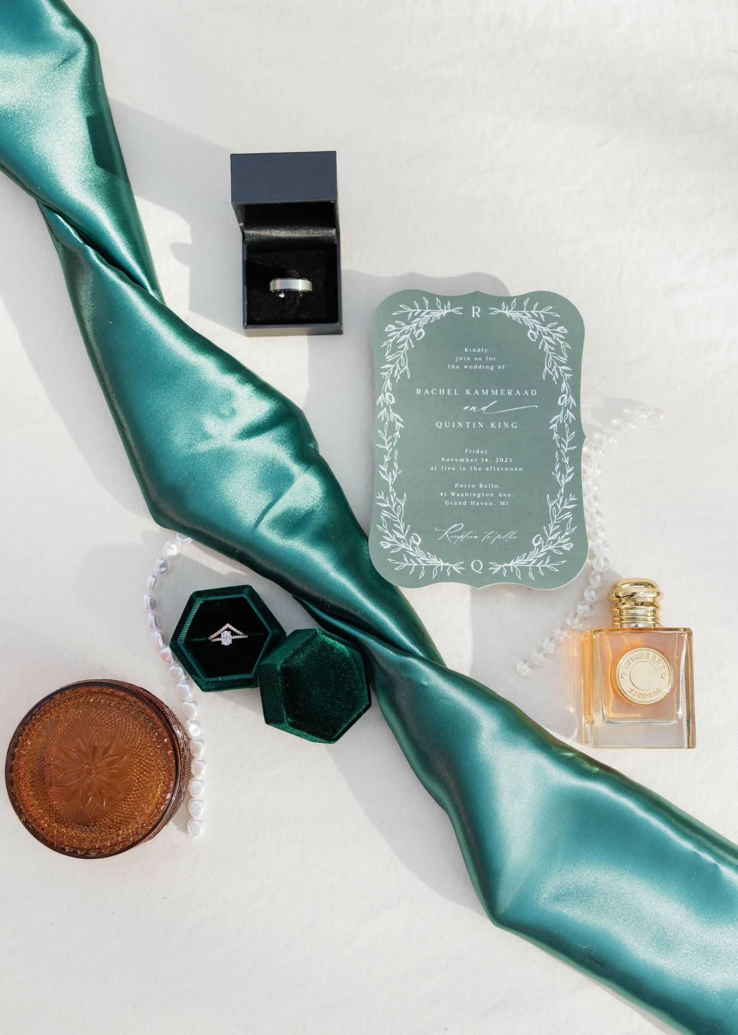 Wedding flat lay featuring emerald green ribbon, ring box, invitation, perfume, and jewelry on white background.