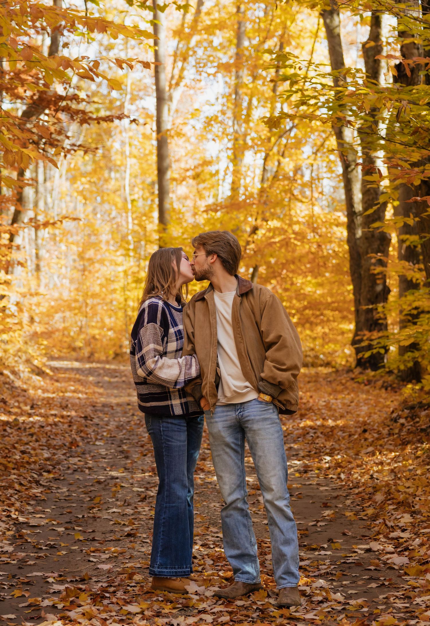 Couple sharing a kiss on a leaf-covered path surrounded by golden autumn trees.