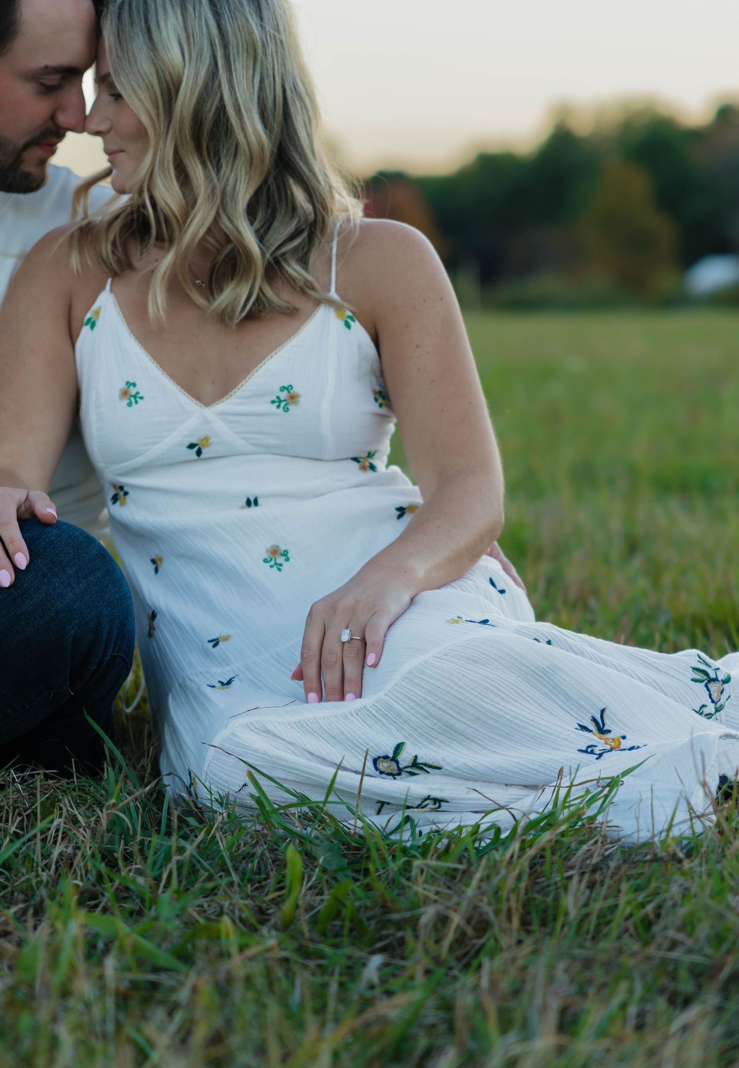 Couple sitting intimately in a grassy field at sunset, woman wearing a white floral embroidered dress.