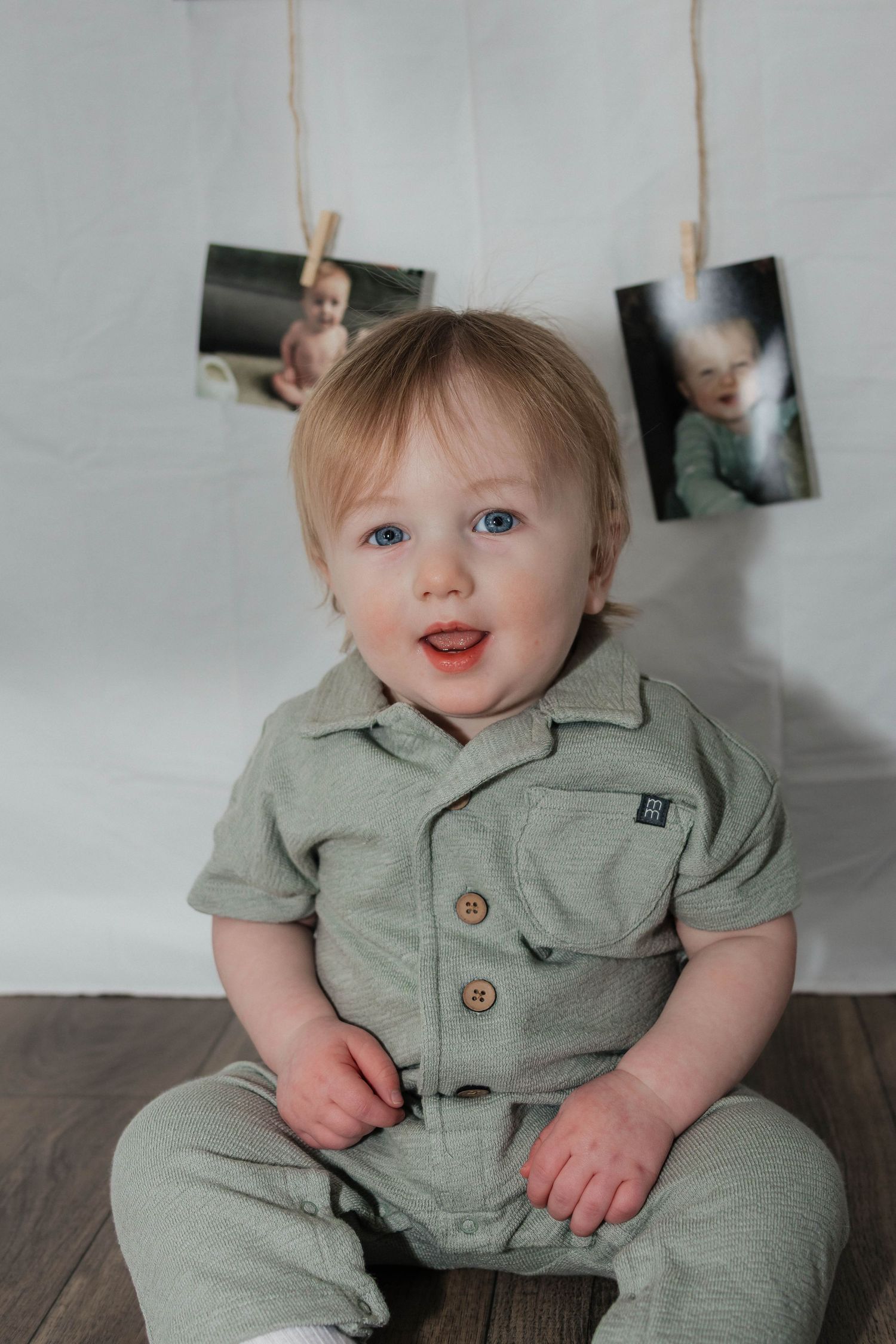 Cute blue-eyed toddler in sage green button romper sitting and smiling in front of hanging photo display.