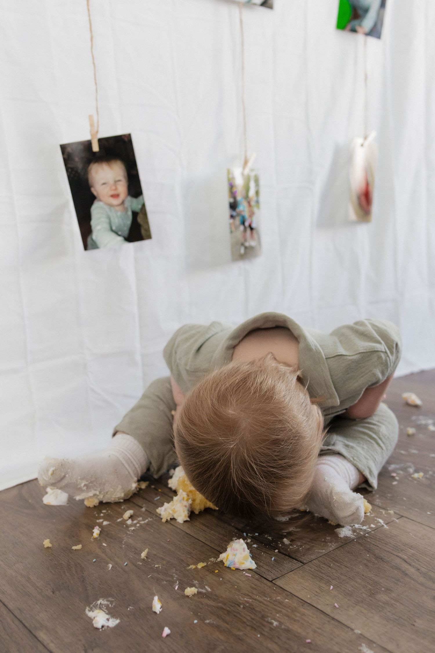 A baby in a green outfit face-down on a wooden floor surrounded by smashed cake crumbs during a cake smash photoshoot.