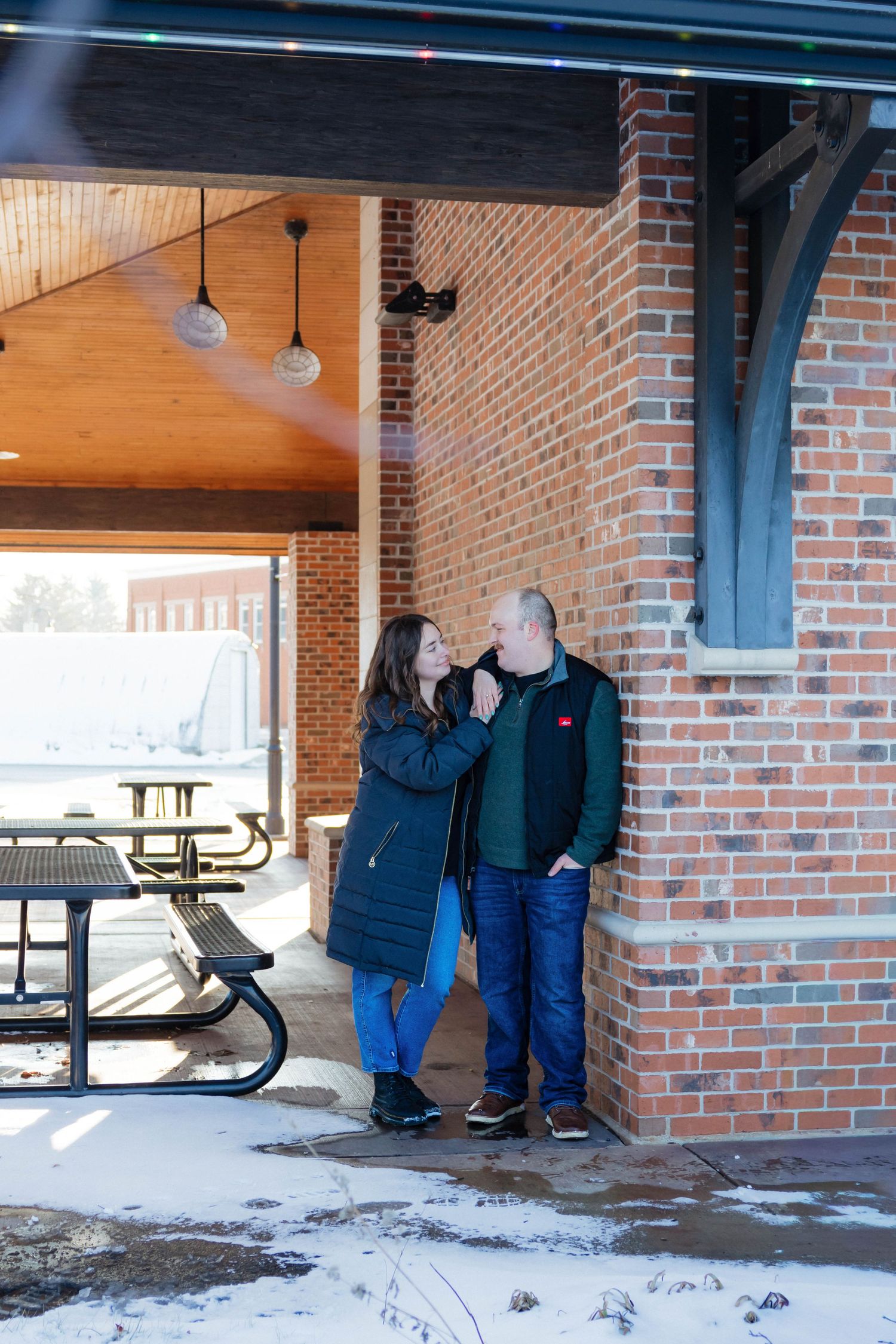 Couple sharing a romantic moment leaning against a brick wall in a snowy outdoor setting.