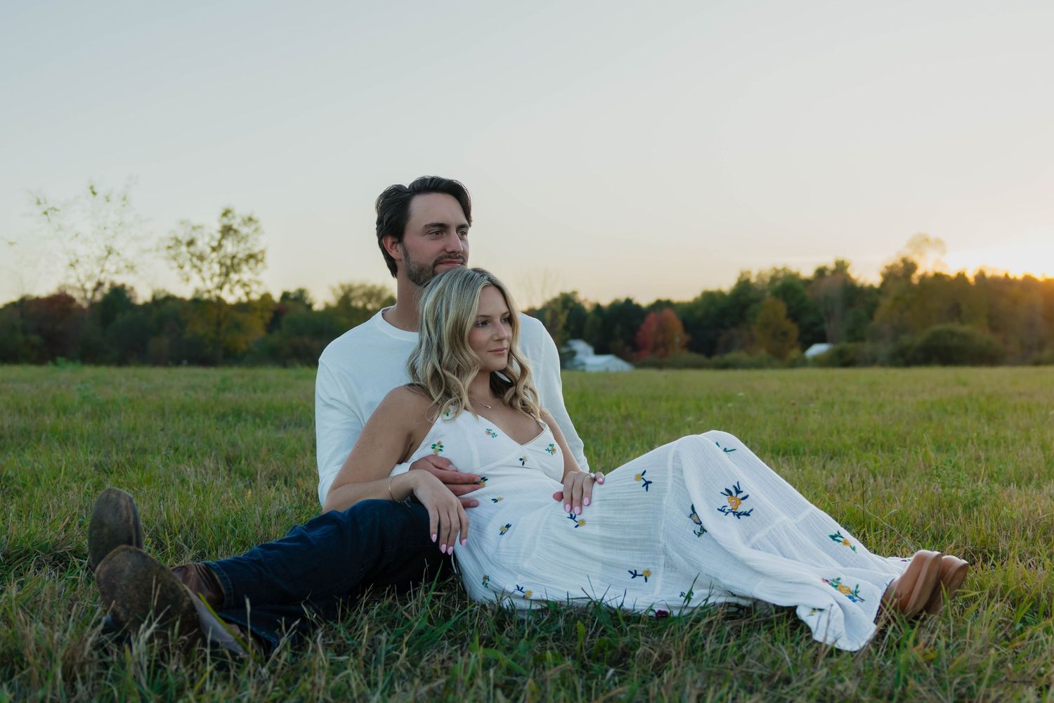 Romantic couple sitting together in an open grassy field during a warm sunset engagement photo shoot.