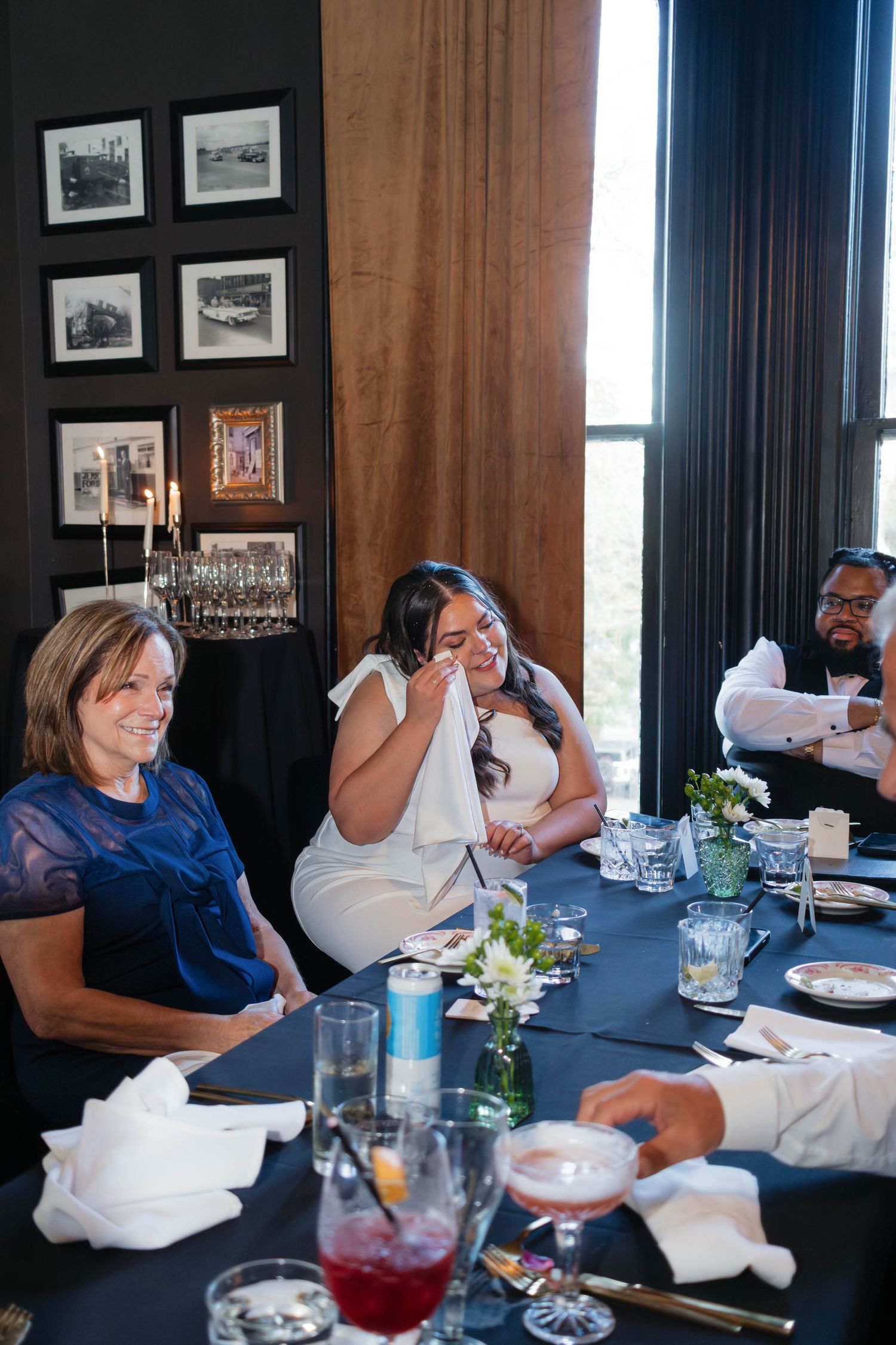 Emotional bride wiping tears at wedding reception dinner table with guests and elegant blue tablecloth.
