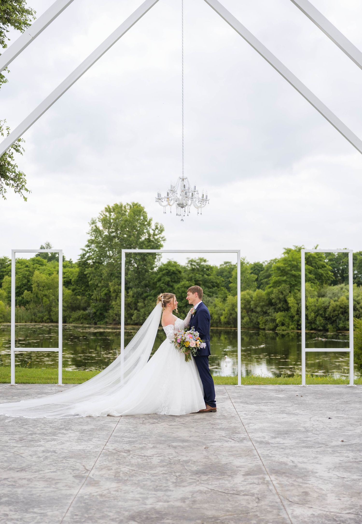 Bride and groom stand beneath a chandelier arch with a scenic pond and lush greenery backdrop at an outdoor wedding.
