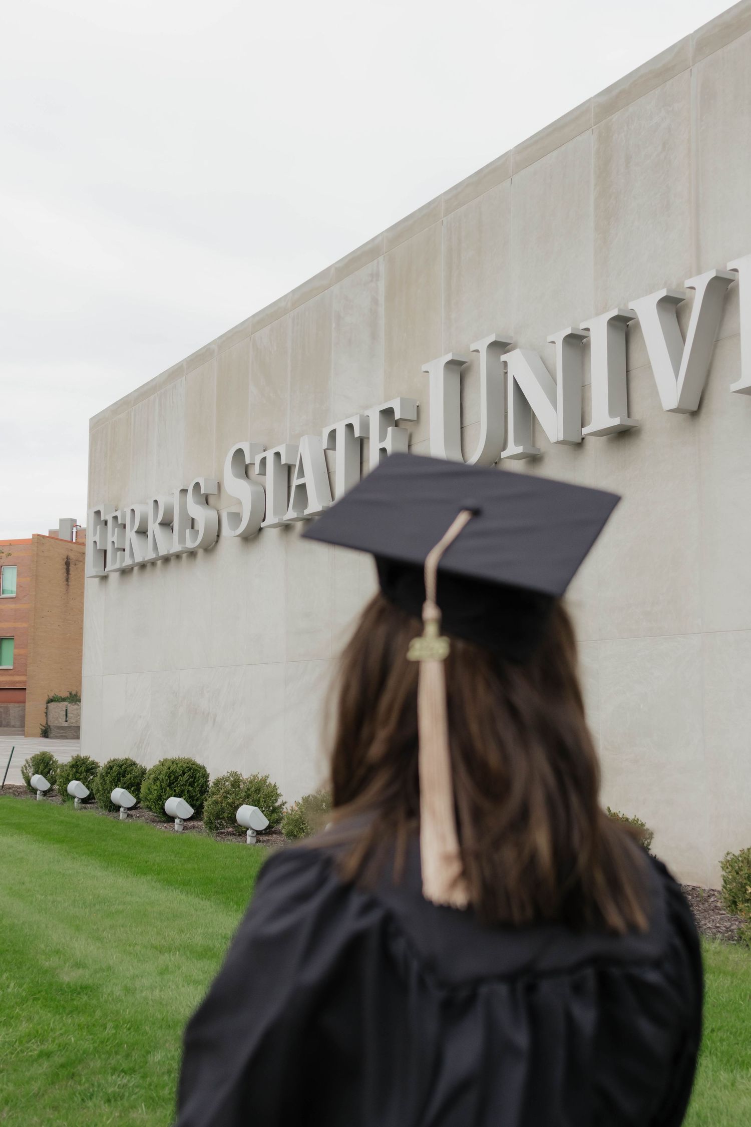Female graduate in cap and gown facing Ferris State University sign on campus.