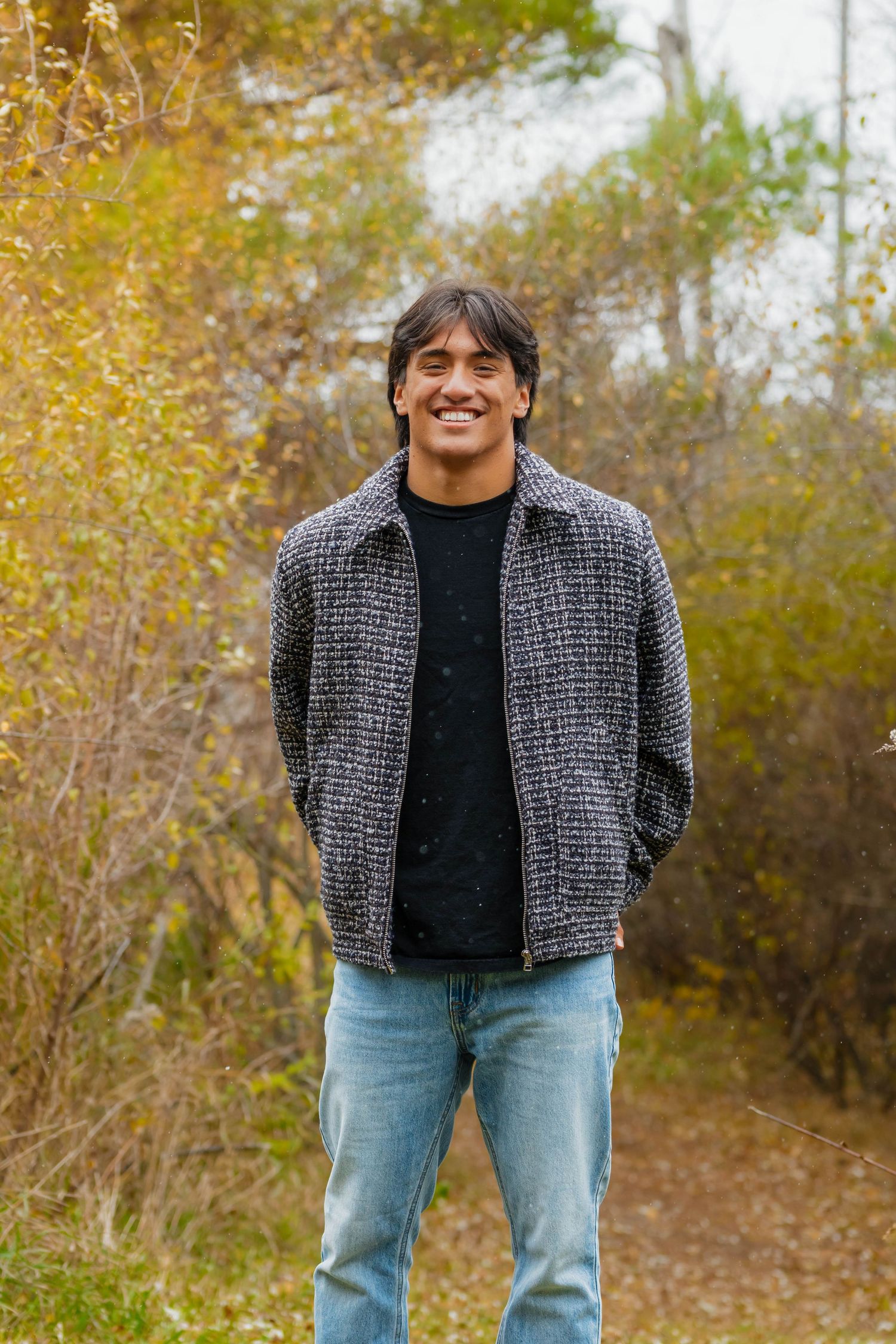 Smiling young man in plaid jacket and jeans stands on autumn nature trail.