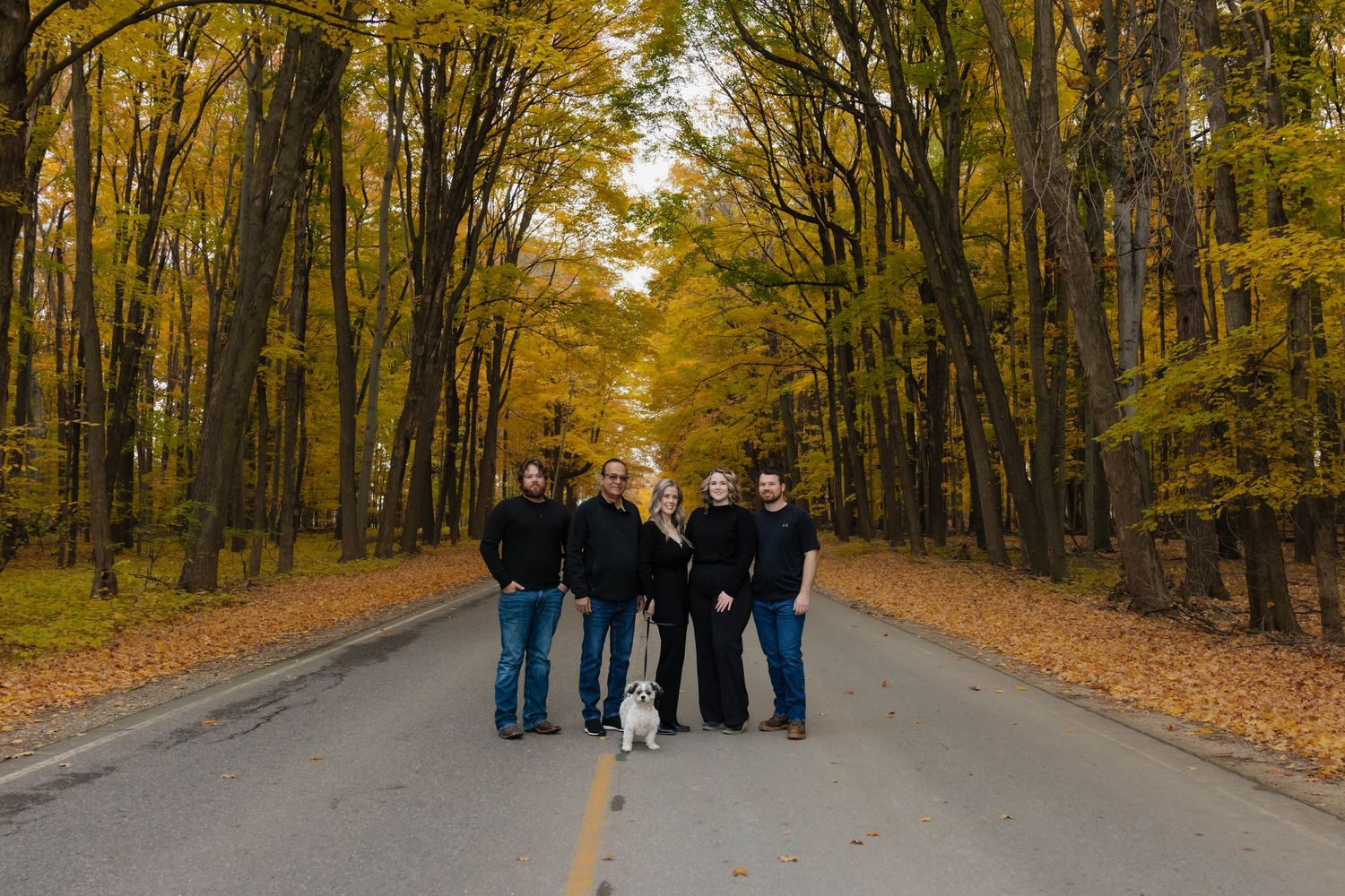 Family group with dog standing on scenic autumn road canopied by vibrant yellow fall trees.