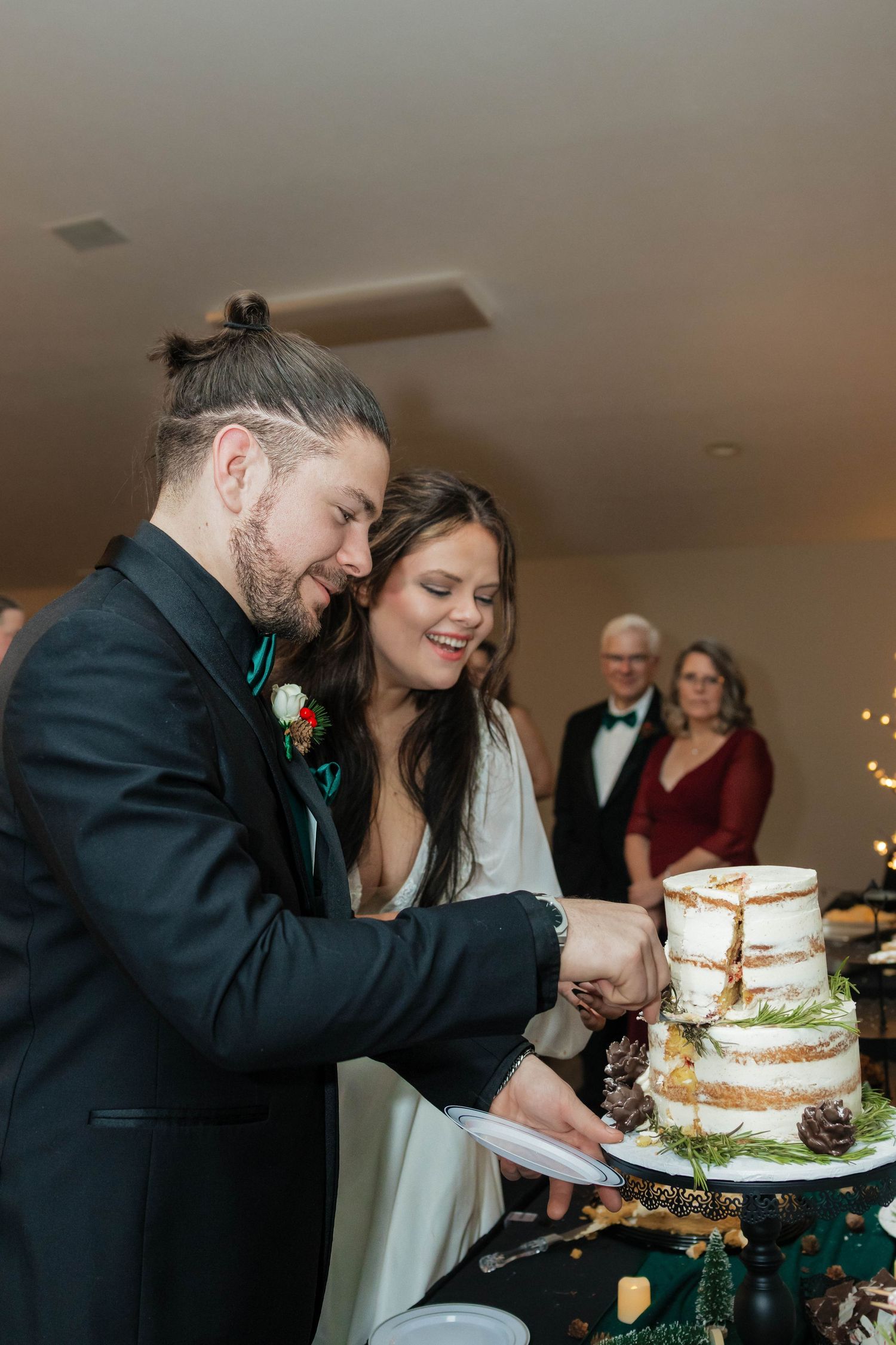 Smiling bride and groom cut rustic naked wedding cake decorated with pine cones and greenery.