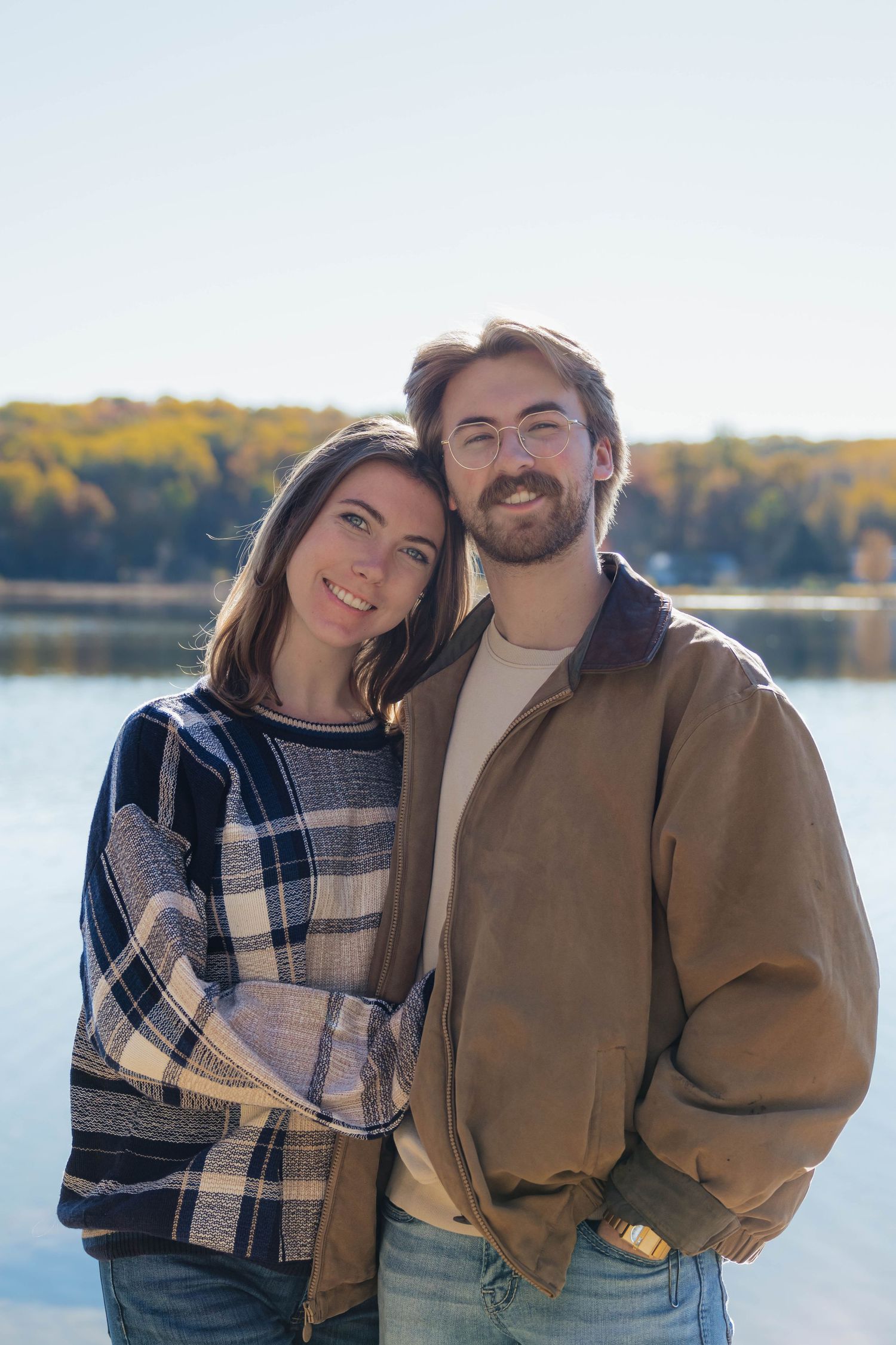 Smiling couple posing together by a sunlit lake surrounded by colorful autumn foliage.