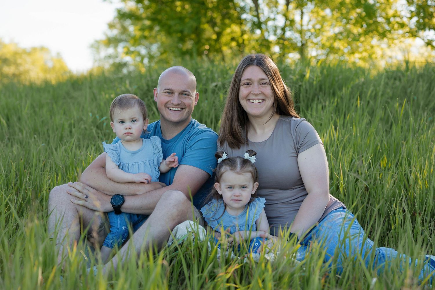 Happy family of four sitting together in tall green grass during golden sunset outdoors.