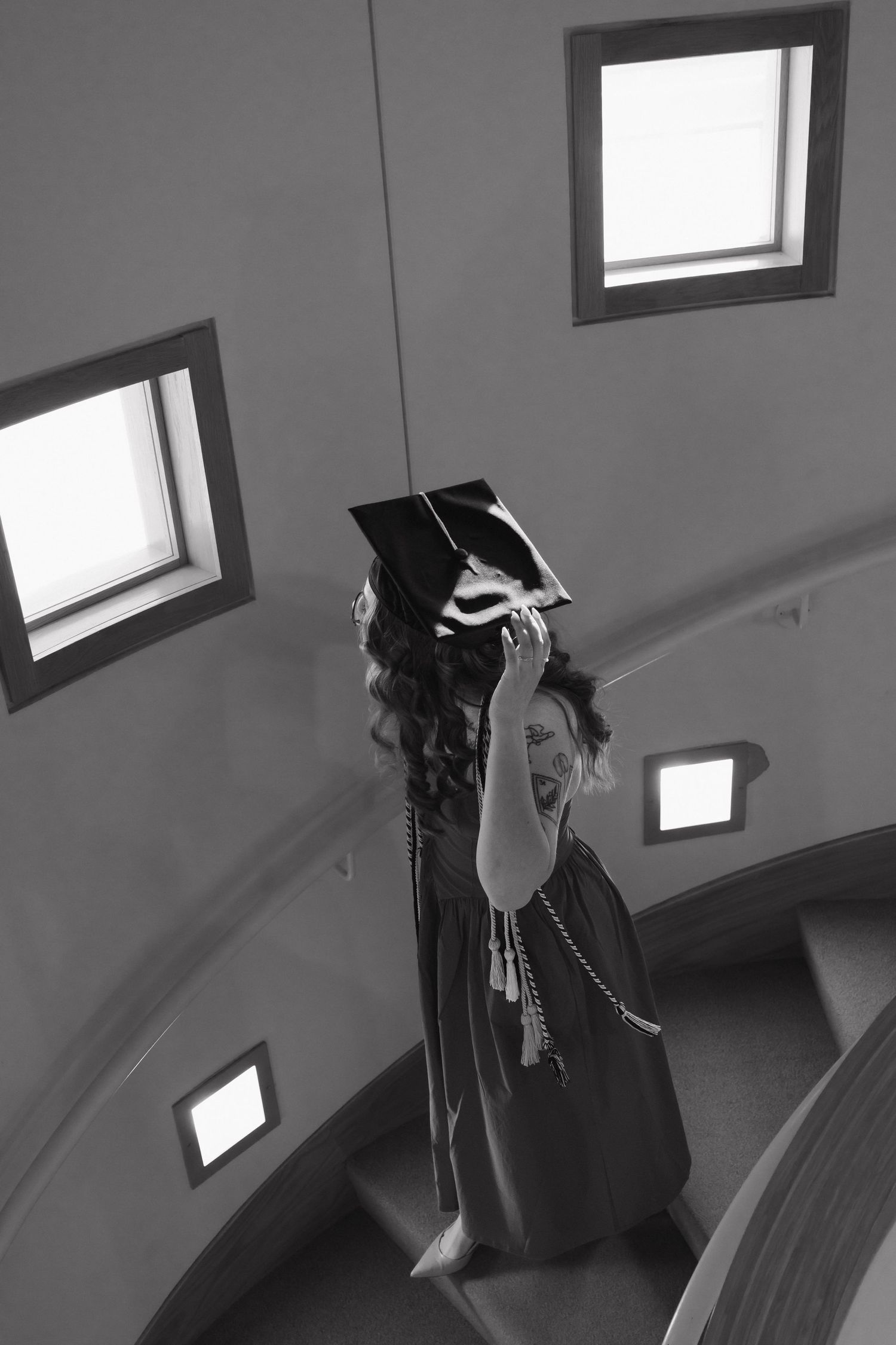 Black and white photo of a graduate in cap and gown raising her cap on a spiral staircase.