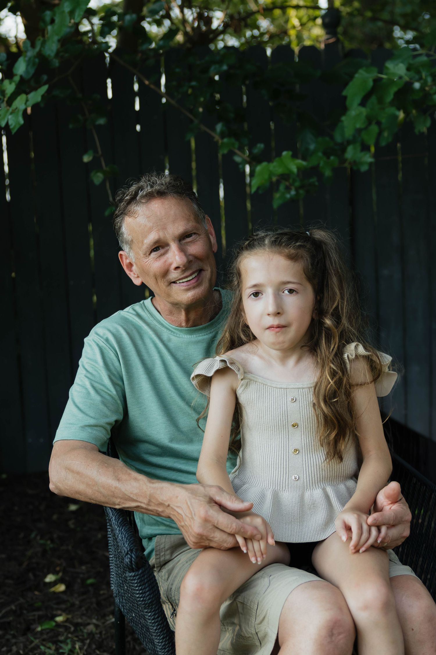 Smiling grandfather sitting outdoors with young granddaughter on his lap near a fence.
