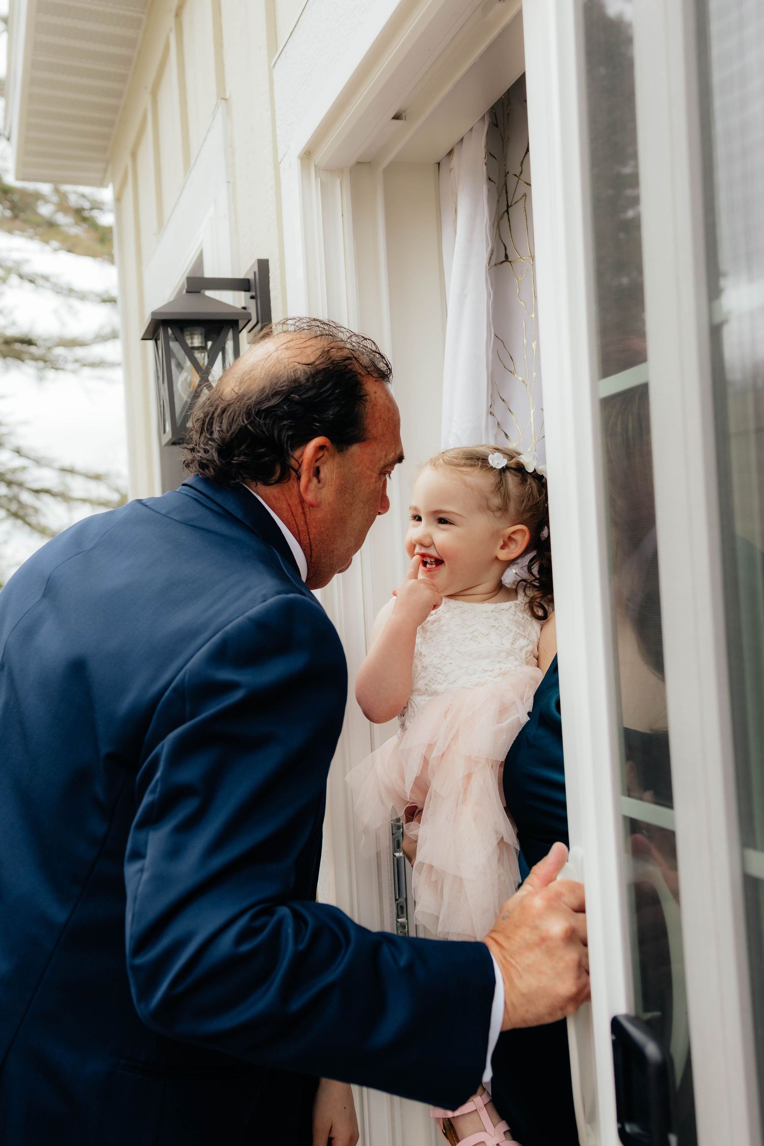 Man in navy suit leaning in to interact with smiling toddler flower girl in pink dress.