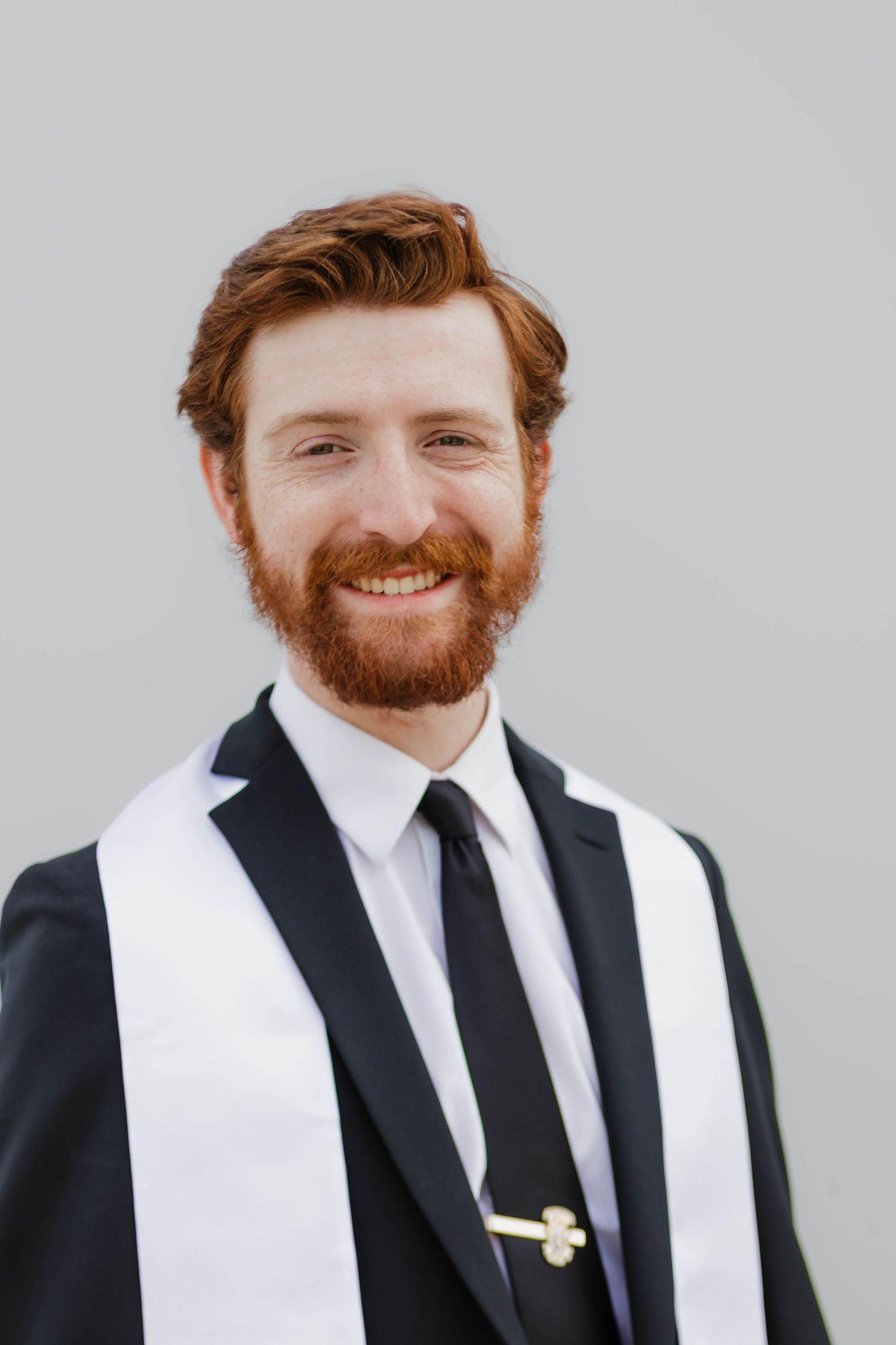 Smiling red-bearded man in black suit with white graduation stole poses for a portrait against a gray background.