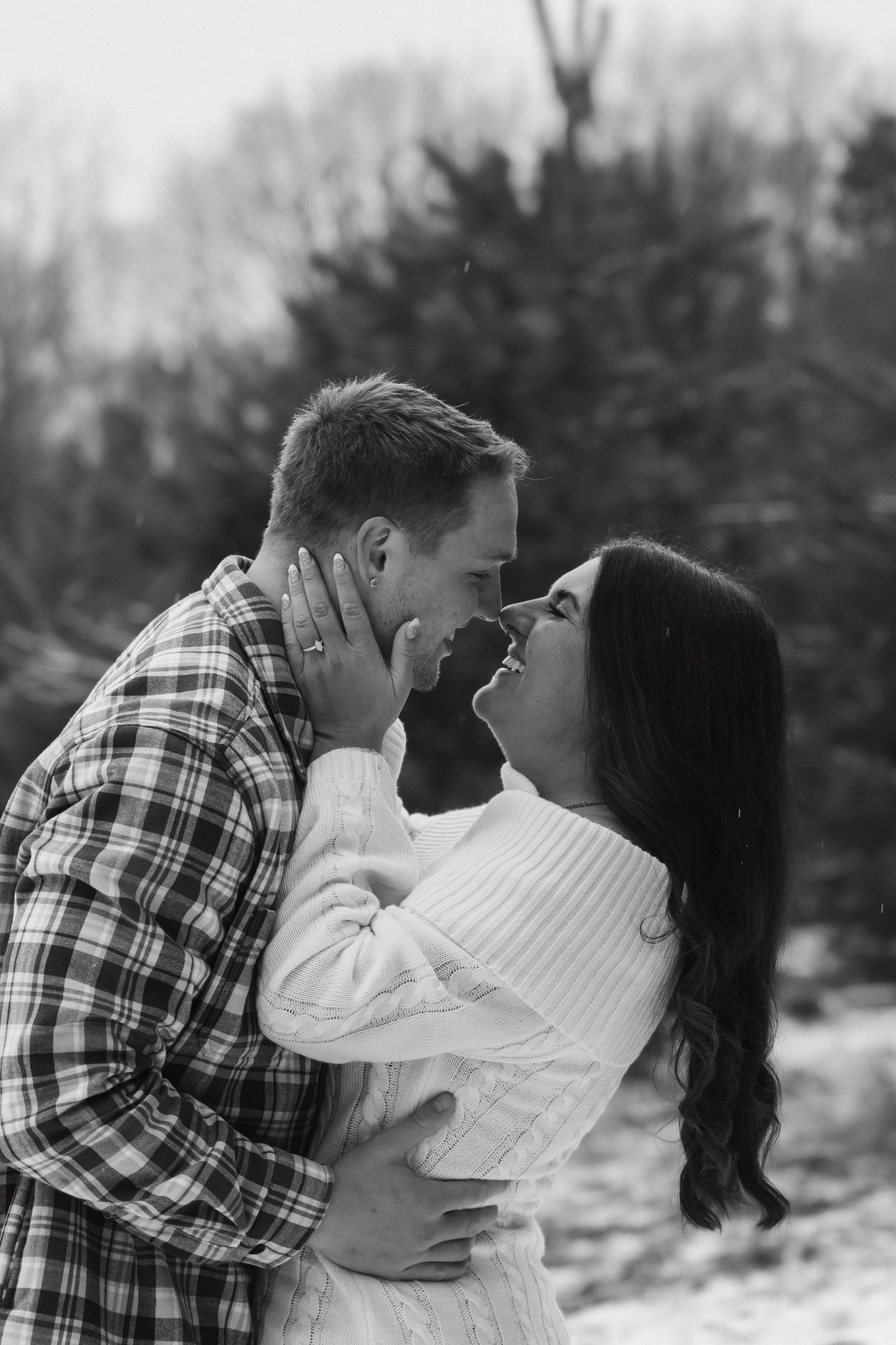 Black and white romantic photo of couple nearly kissing in snowy pine forest engagement session.