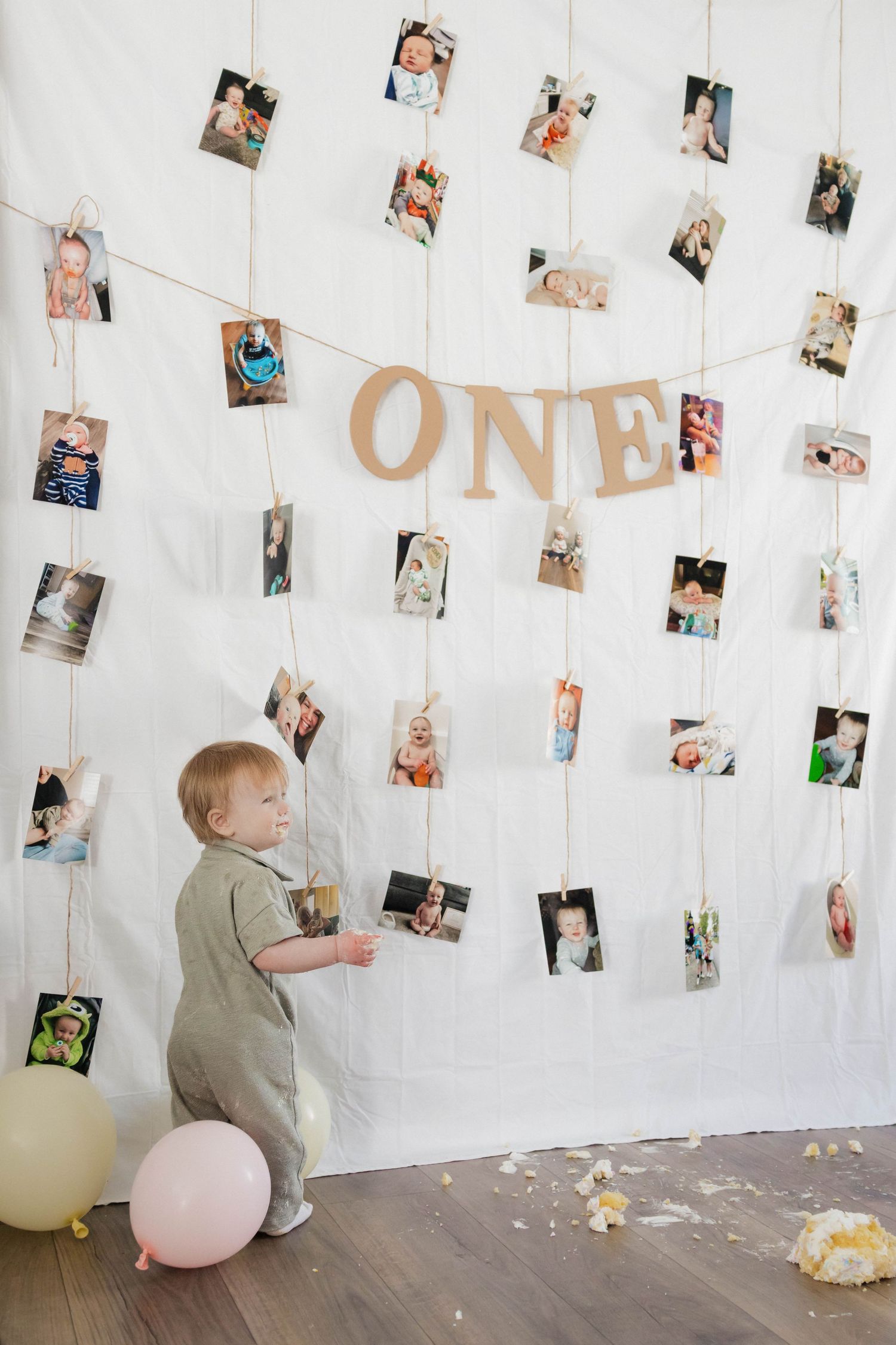 One-year-old boy stands before large photo display wall with ONE banner and balloons.