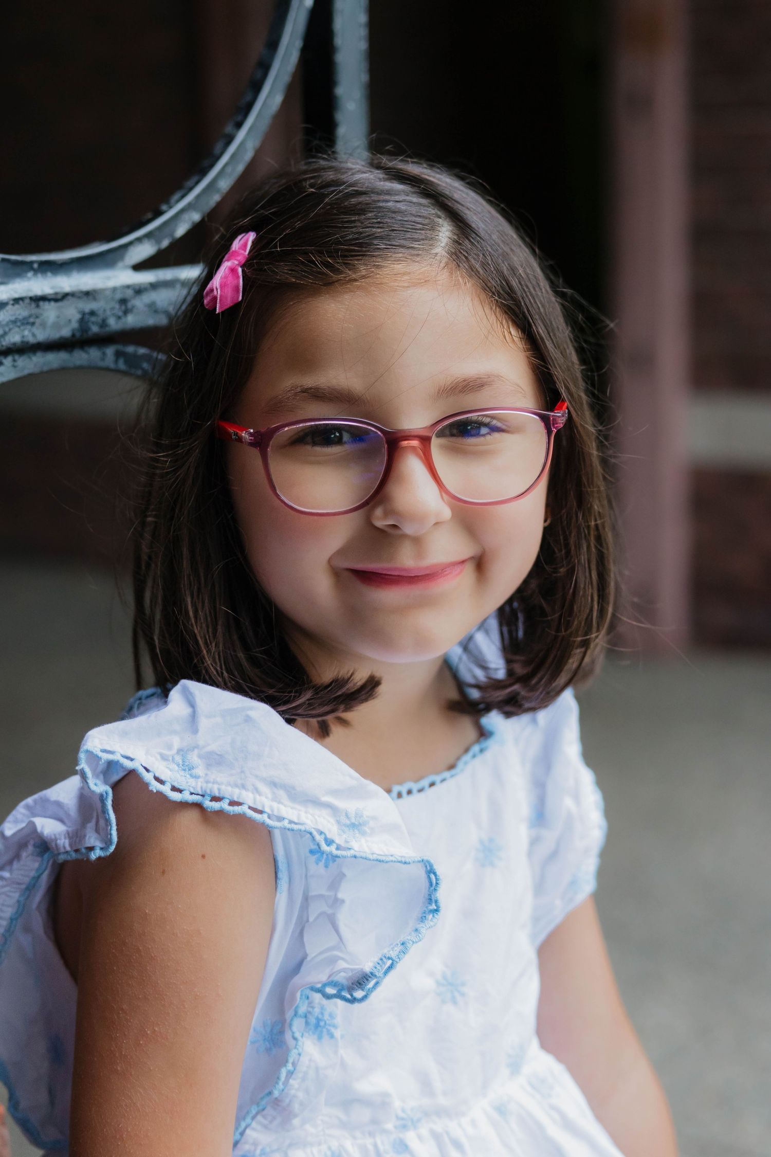 Close-up portrait of a young girl with red glasses and a pink hair clip wearing a white dress outdoors.