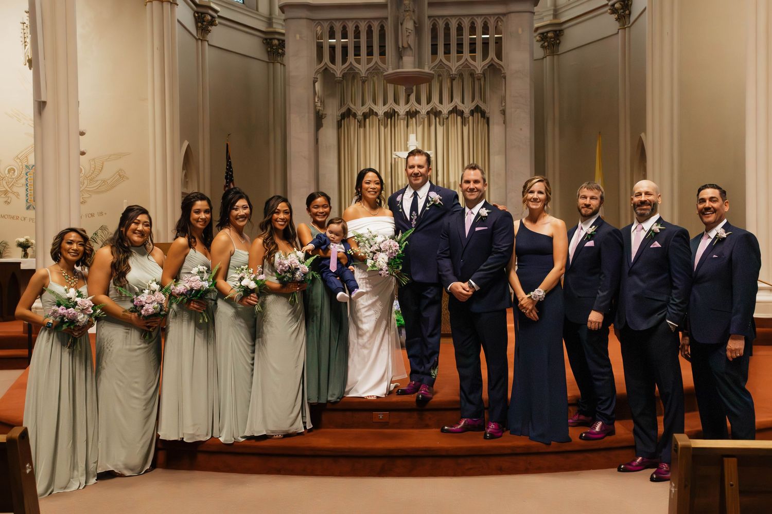 Wedding party posing in a church with bridesmaids in sage green gowns and groomsmen in navy suits.
