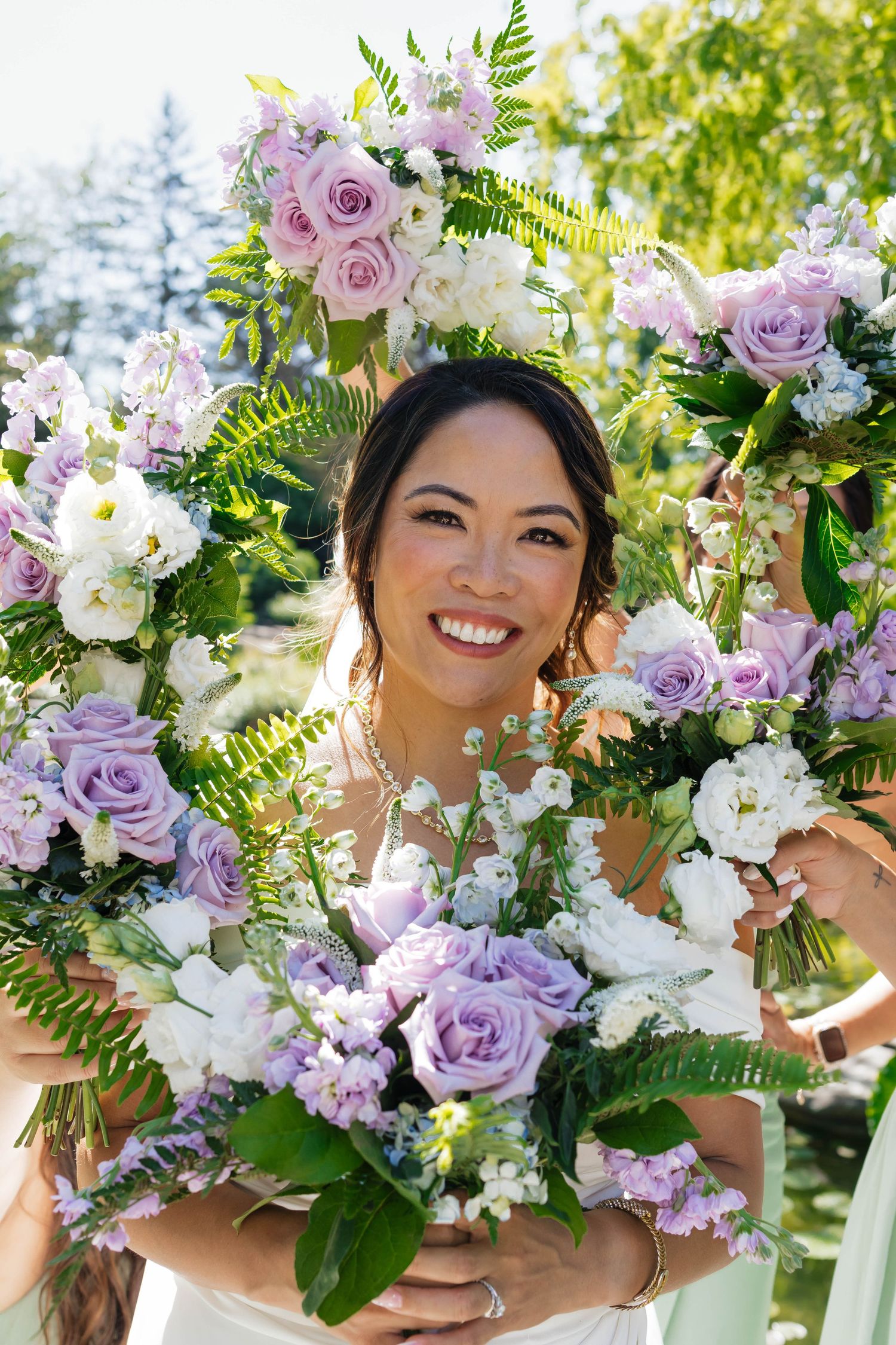 Radiant bride smiles while framed by lavender and white rose bouquets held by bridesmaids in sunny garden setting.
