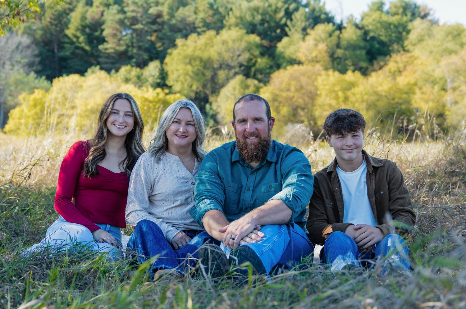 Family of four sitting in a grassy field surrounded by autumn trees, smiling for a portrait.