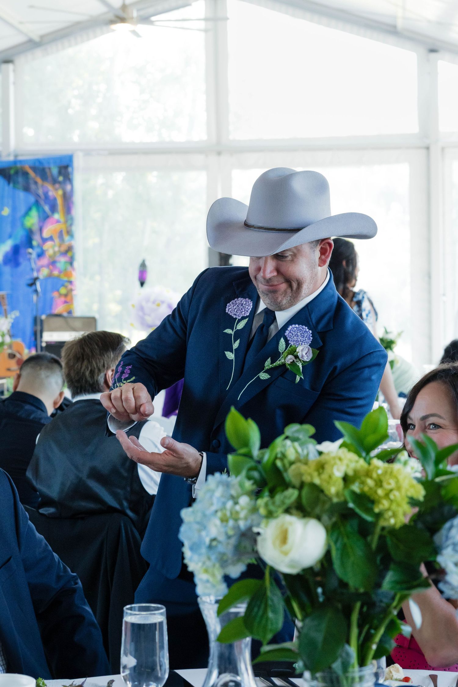 Groom in navy suit and gray cowboy hat smiles while interacting with guests at wedding reception.