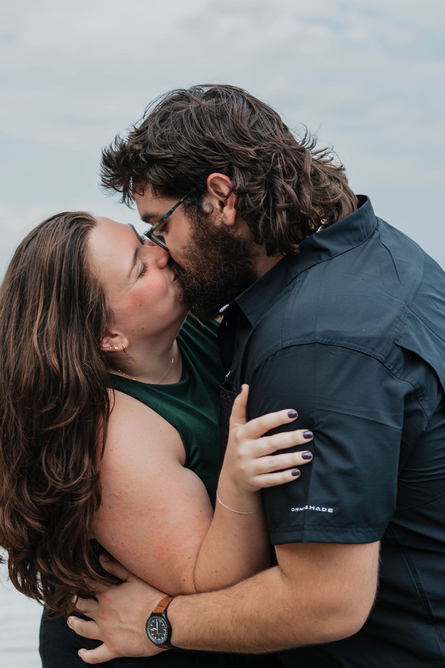 A couple shares a romantic kiss outdoors near water, with the woman embracing the man tenderly.