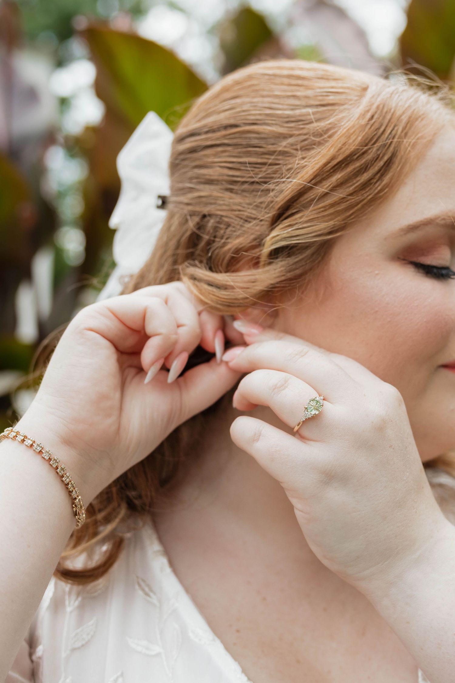 Bride with red hair adjusting her earring, wearing a diamond engagement ring and gold bracelet on her wedding day.