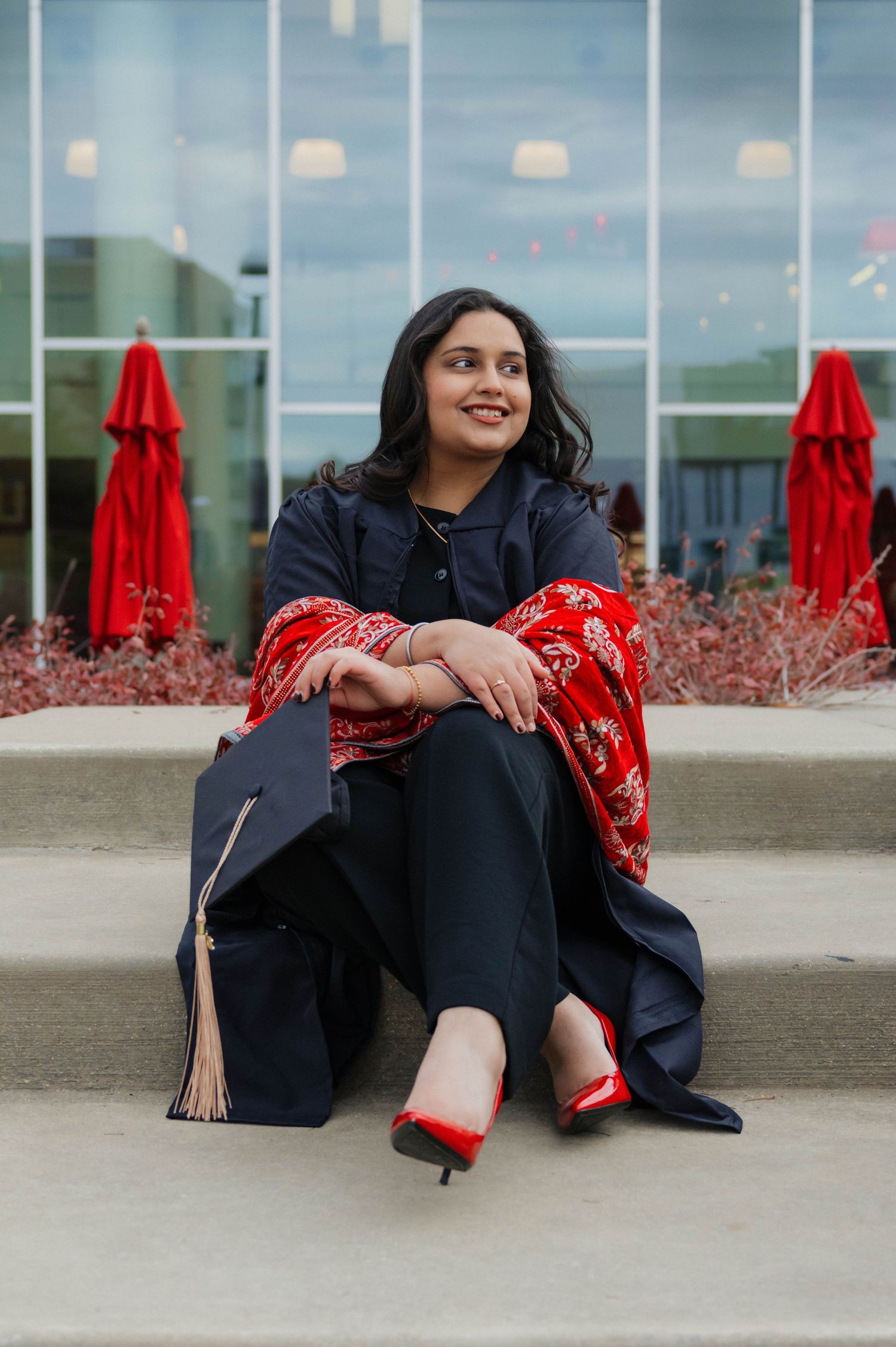 Young woman in graduation cap and red dupatta sits on steps smiling, celebrating academic achievement.
