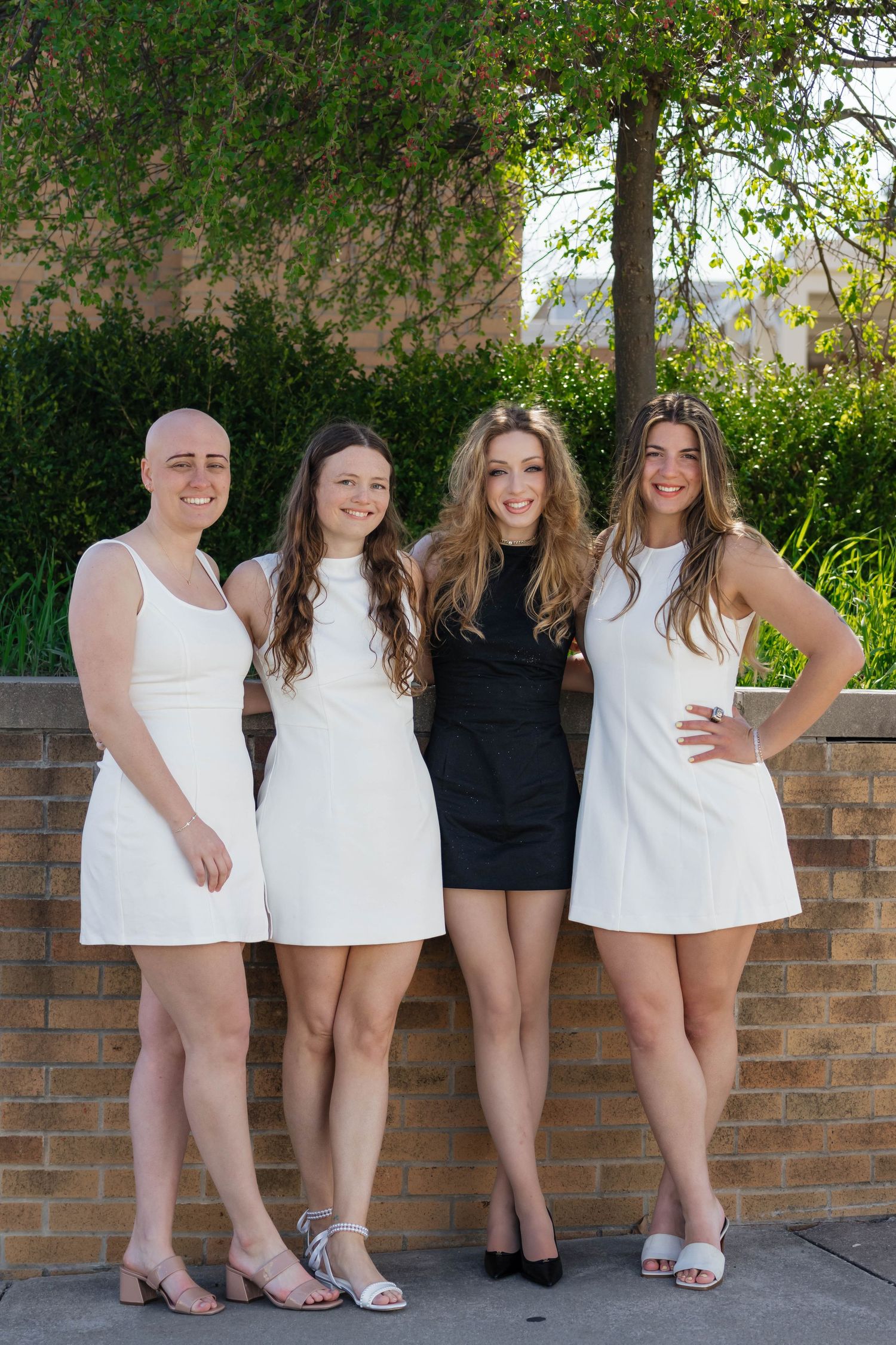 Four young women in white and black mini dresses pose together outdoors near a brick wall.