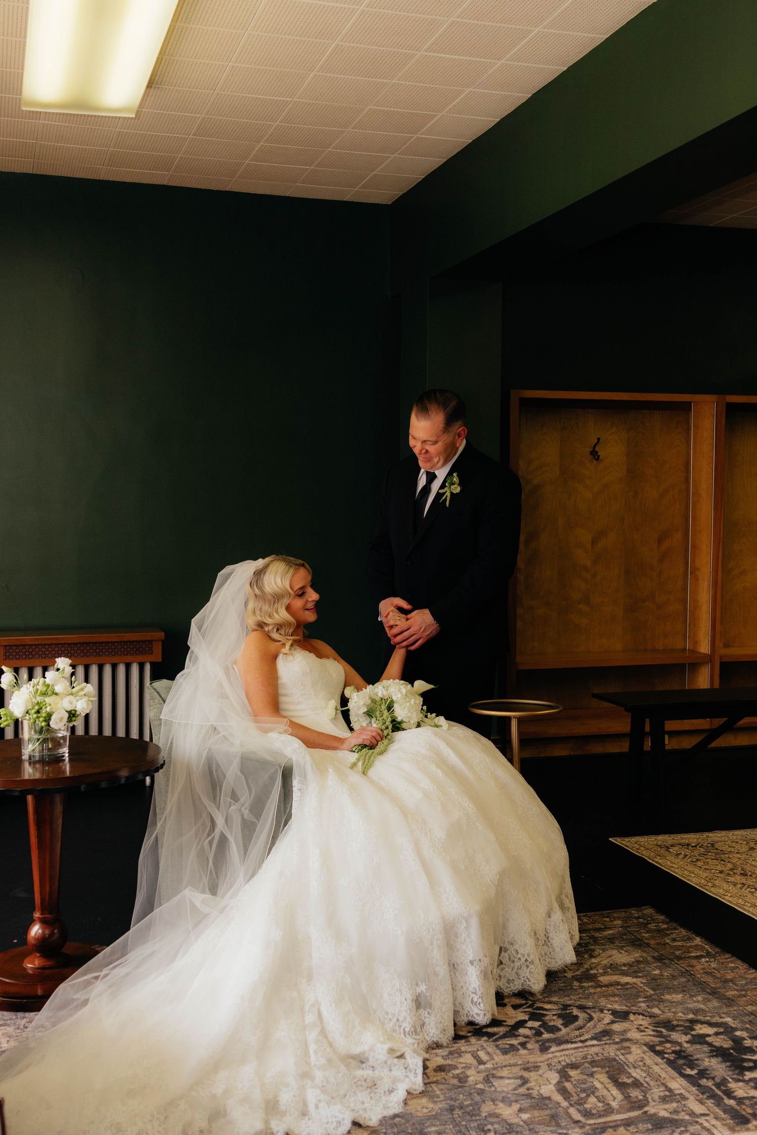Bride in lace wedding gown seated with father holding hands in dark-walled room before ceremony.
