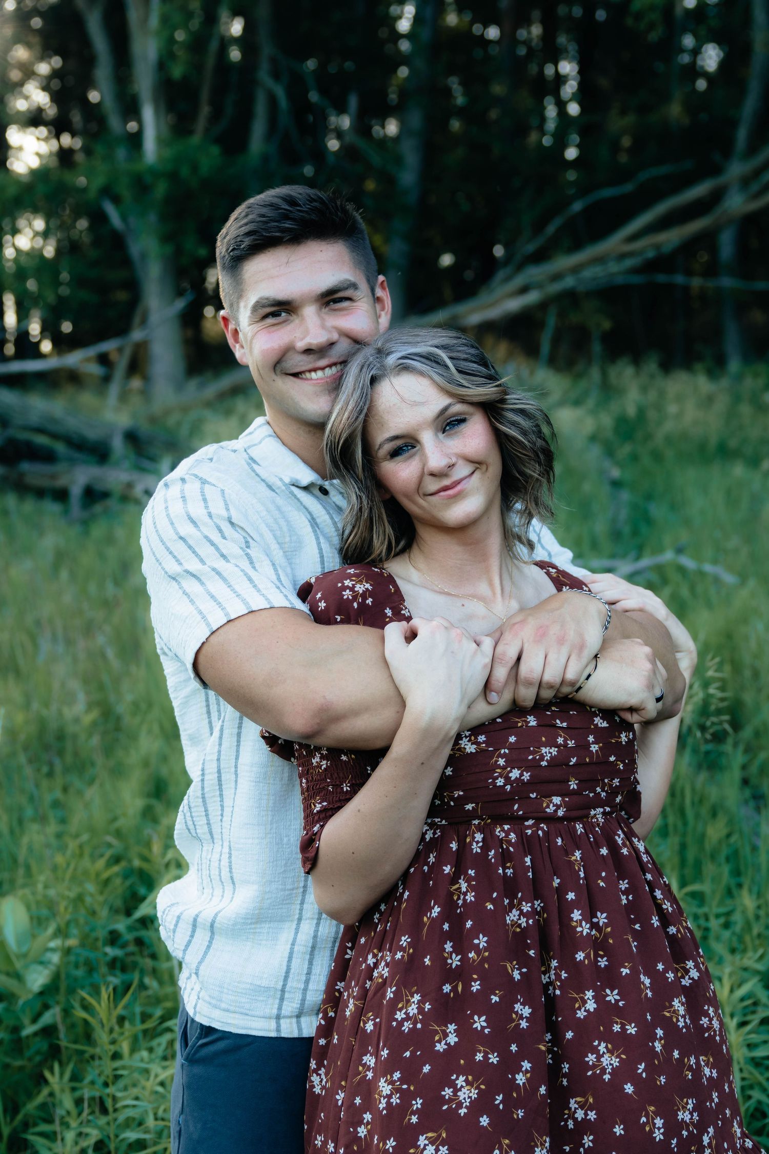 Smiling couple embracing in golden hour light in a forest meadow, man hugging woman in burgundy dress.